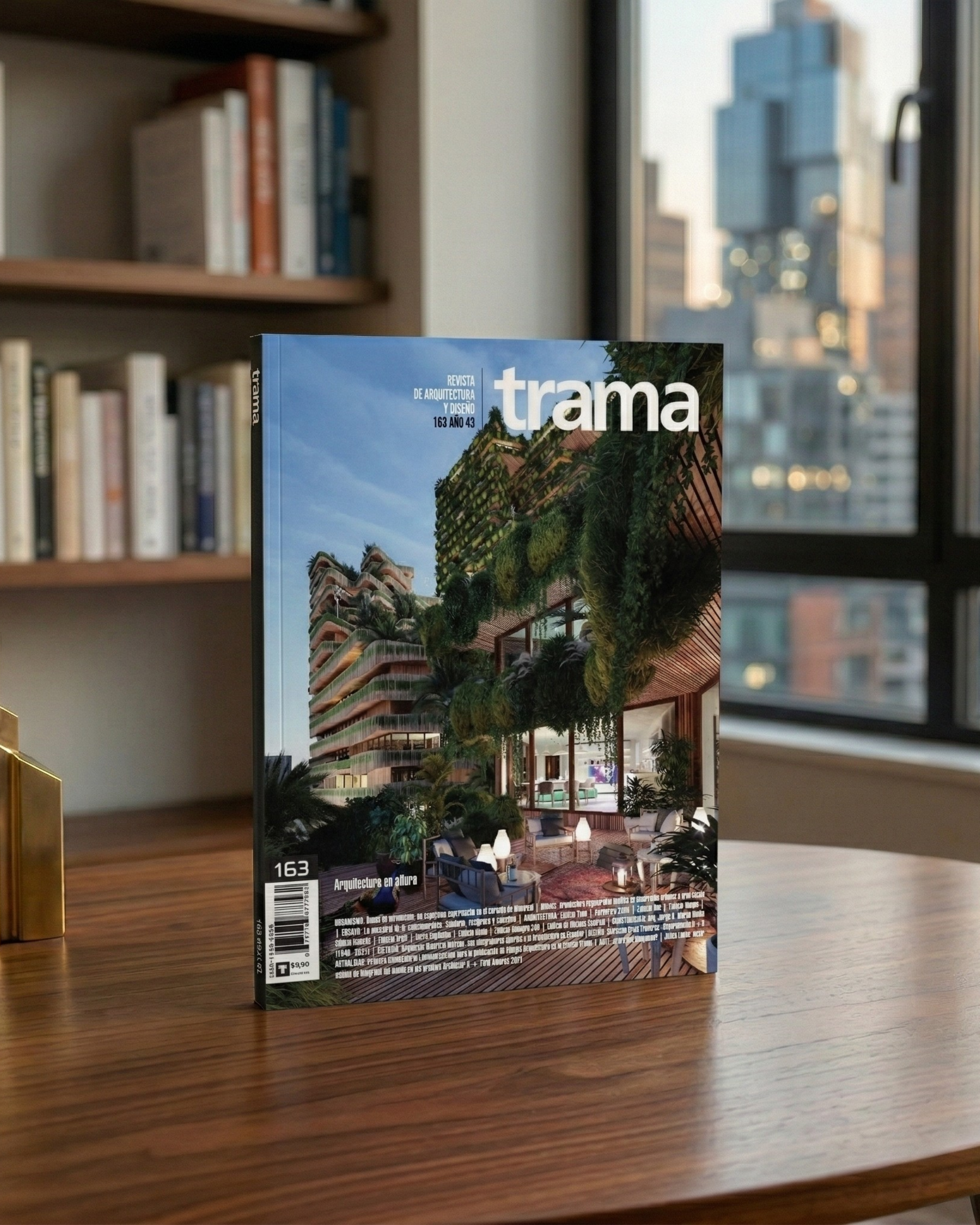 Revista de arquitectura y diseño titulada 'trama', con una portada que muestra un edificio moderno con áreas verdes en su fachada y un espacio de terraza con mobiliario en un entorno urbano, en una mesa de madera en una oficina con estanterías y ventanas que muestran edificios en la ciudad.