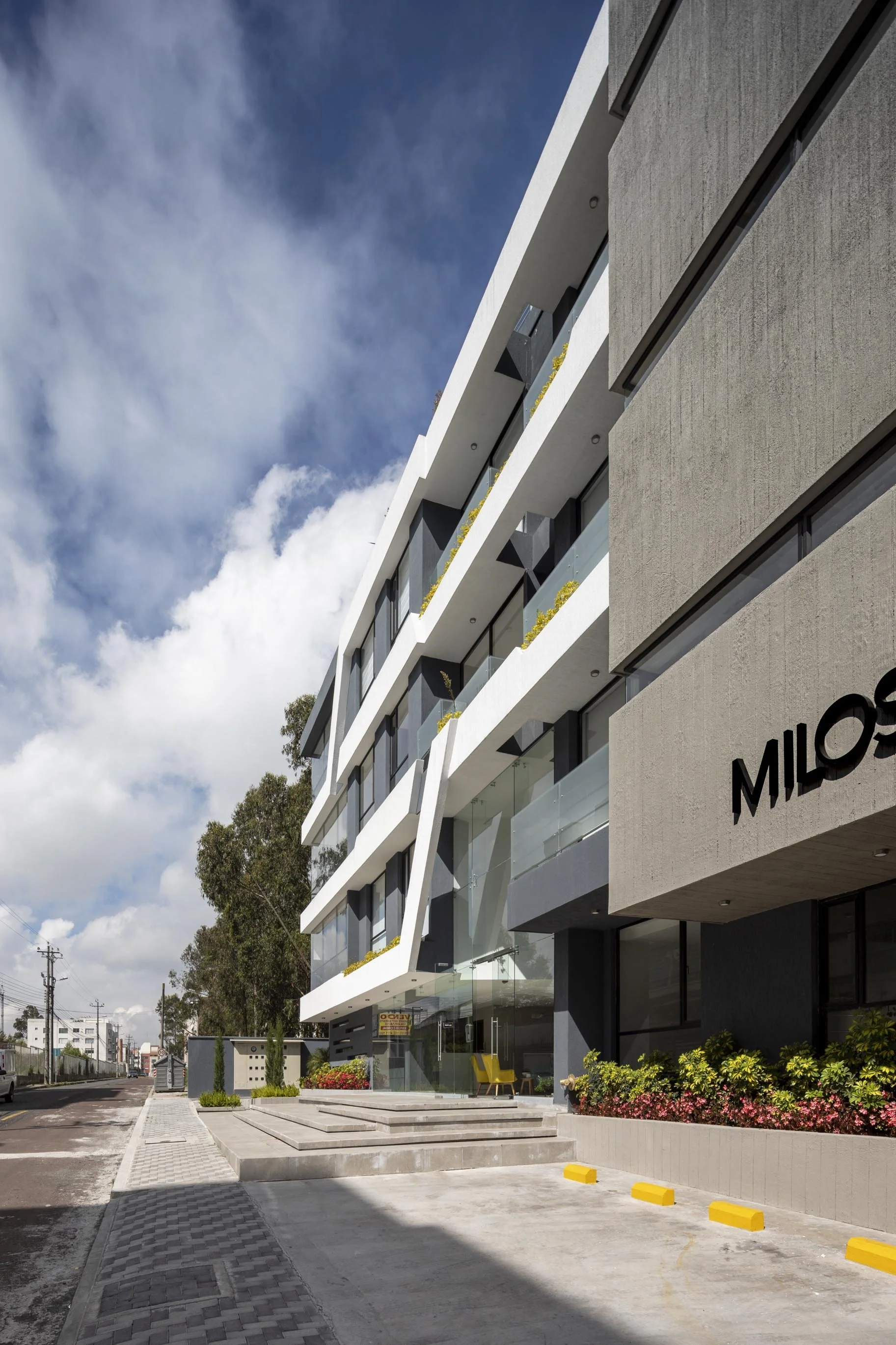 Edificio moderno de apartamentos con fachada de cemento y cristales, con plantas en los balcones y un acceso elevado con escaleras, en una calle con árboles y cielo nublado.