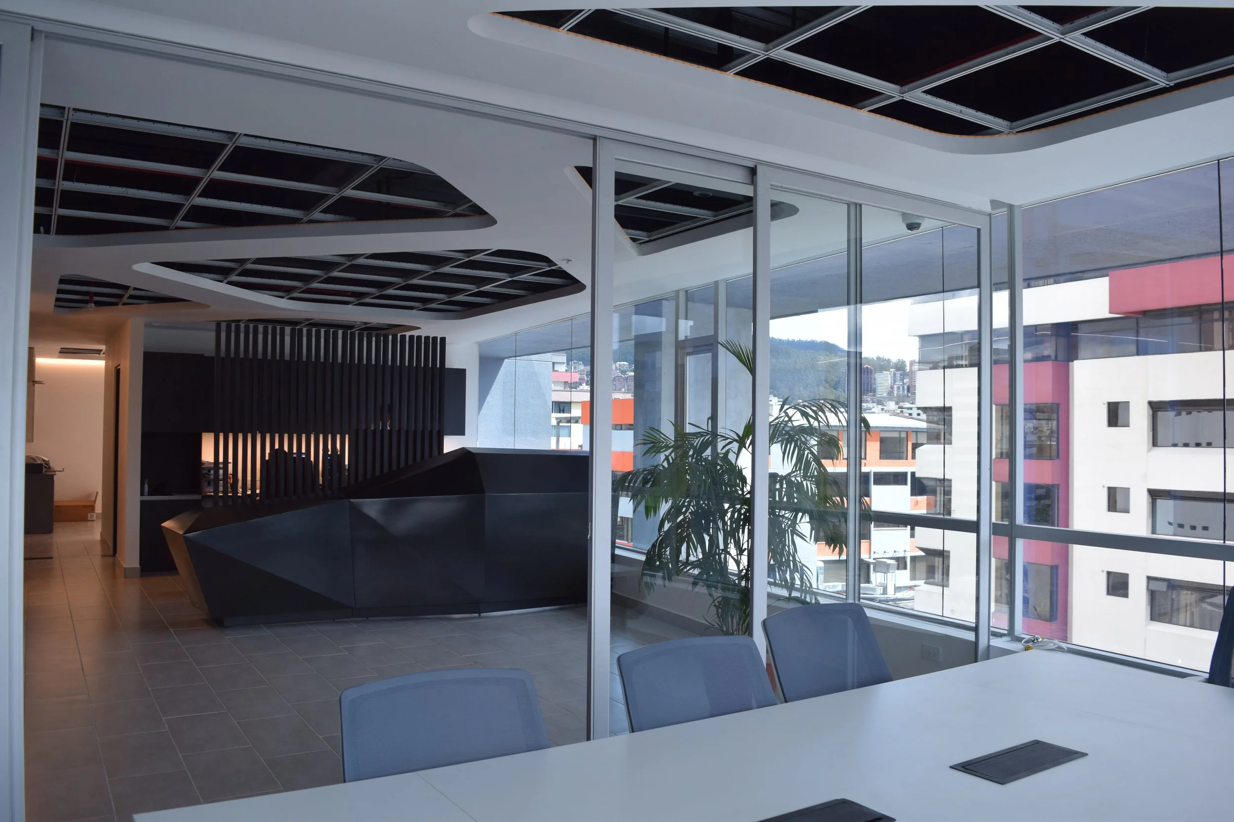 Vista de una oficina moderna con paredes de vidrio, mesas y sillas, y una vista de edificios y montañas en el exterior.