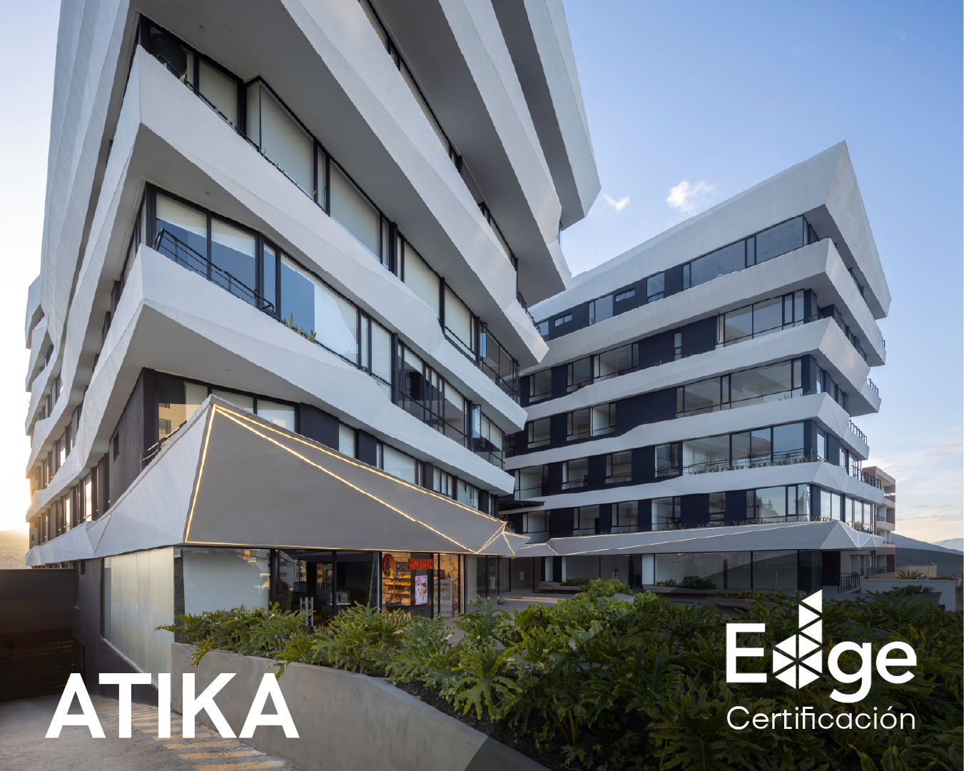 Edificio moderno de varias plantas con balcones y ventanas de vidrio, con áreas verdes en la base, durante el atardecer con cielo despejado.