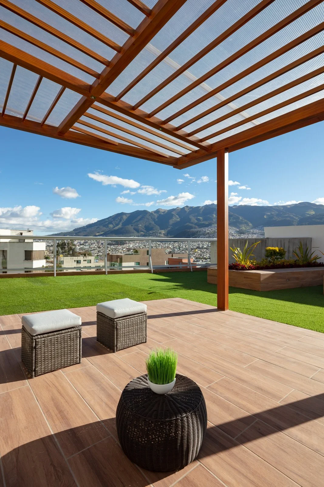 Terraza con piso de madera, sombra de pergola de madera, vista a montañas y ciudad, plantas en macetas, sillas de mimbre con cojines blancos, cenicero decorativo con planta artificial, cielo azul con algunas nubes.