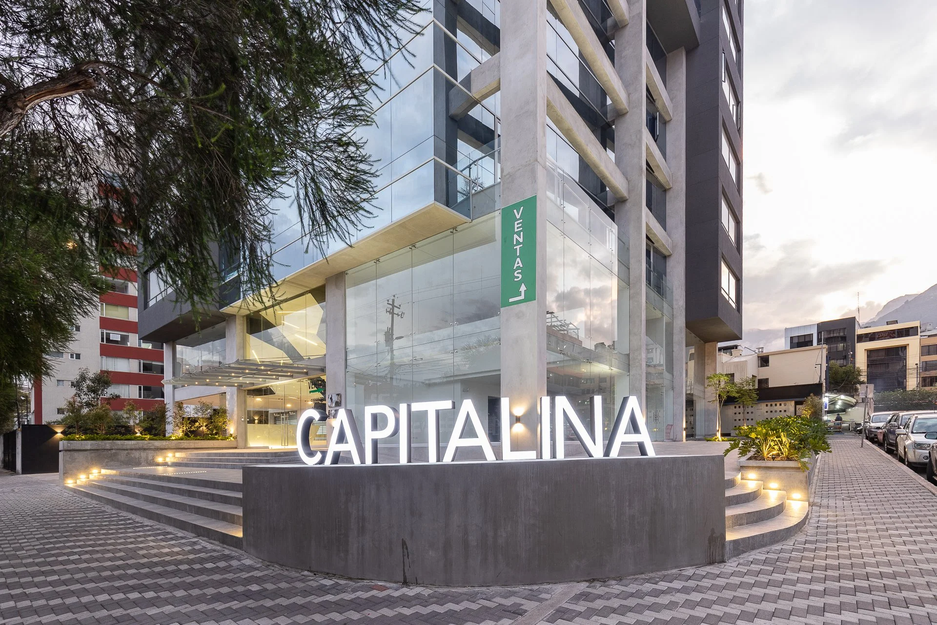 Edificio moderno con letrero de neón que dice 'CAPITÁLINA' en la entrada, ubicado en una calle con árboles y vehículos estacionados.