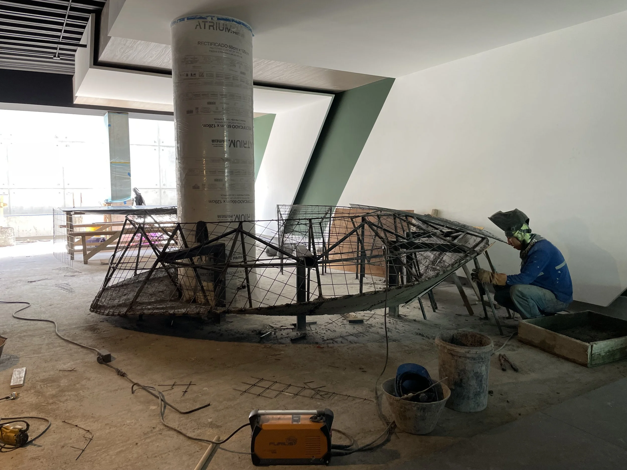 Trabajador en obra en interior, soldando estructura de metal en el suelo, rodeado de herramientas y materiales de construcción.