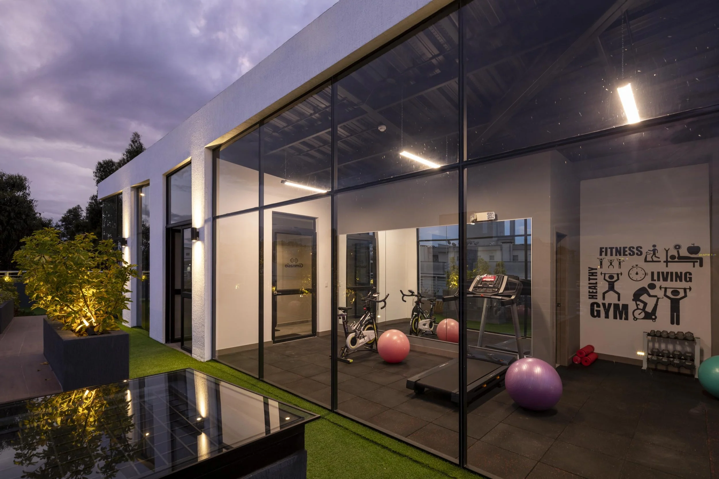 Vista exterior de un gimnasio moderno con paredes de cristal, equipado con bicicletas estáticas, balón de ejercicio, y máquinas de entrenamiento, con un mural que dice "Fitness, Healthy, Gym, Living" en la pared interior.
