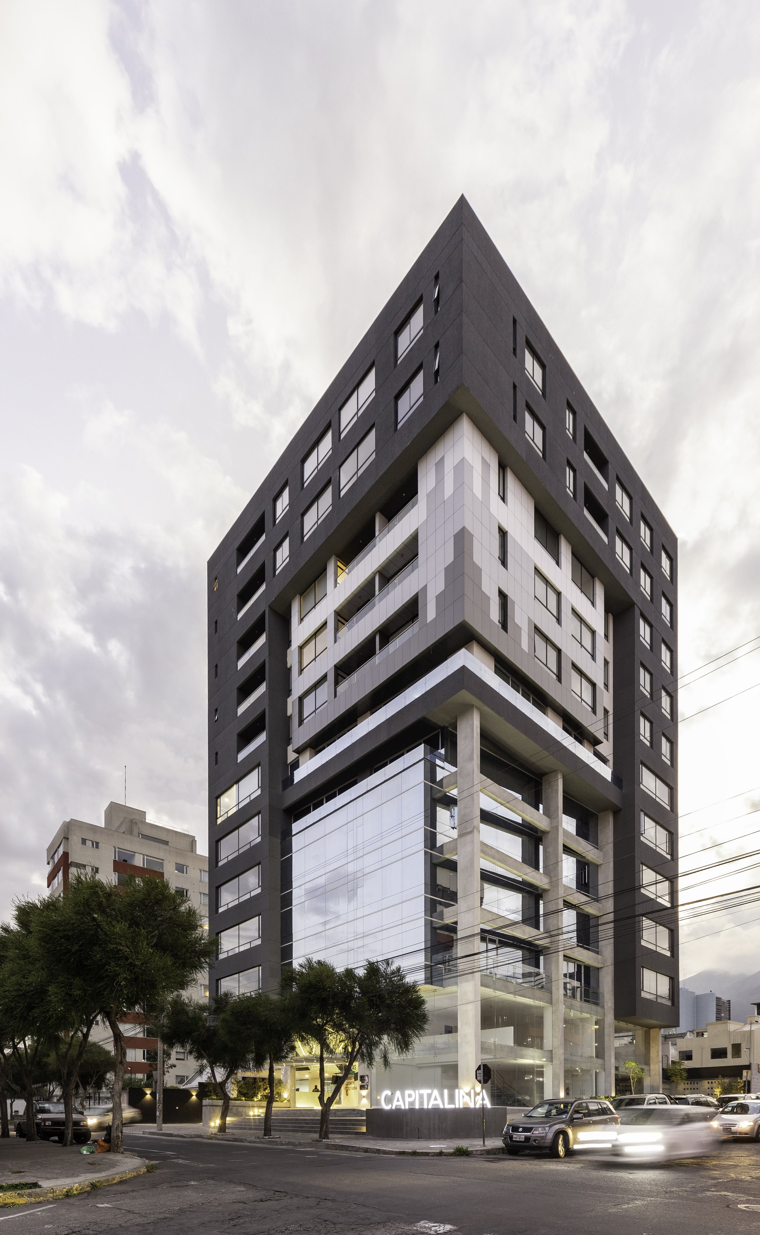 Edificio moderno de varios pisos con fachada de vidrio y estructura negra, con árboles y autos en la calle en primer plano, y cielo nublado en el fondo.