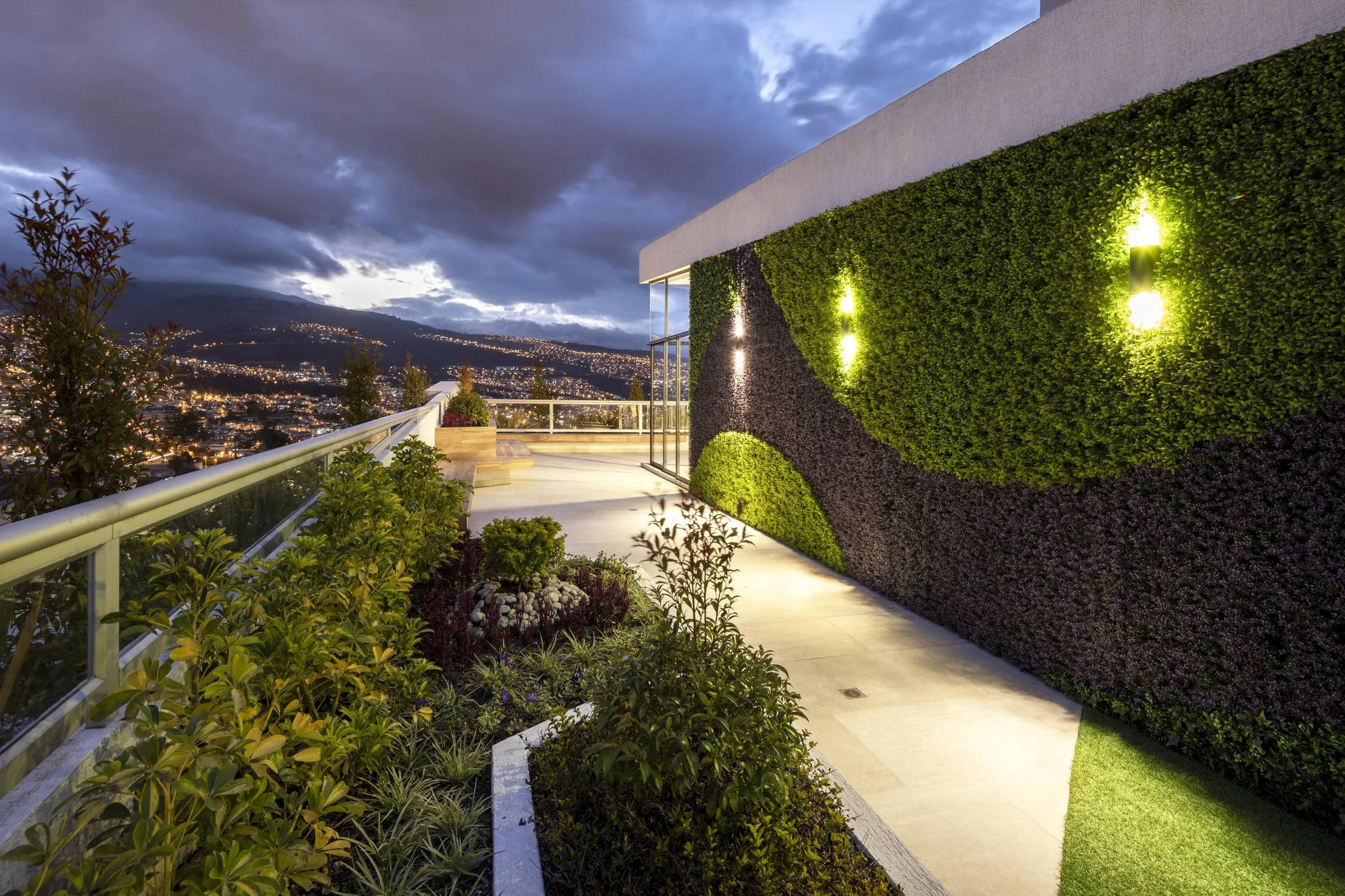 Terraza moderna en la ciudad con jardines y muro vegetal iluminado, vista nocturna de la ciudad en el fondo.
