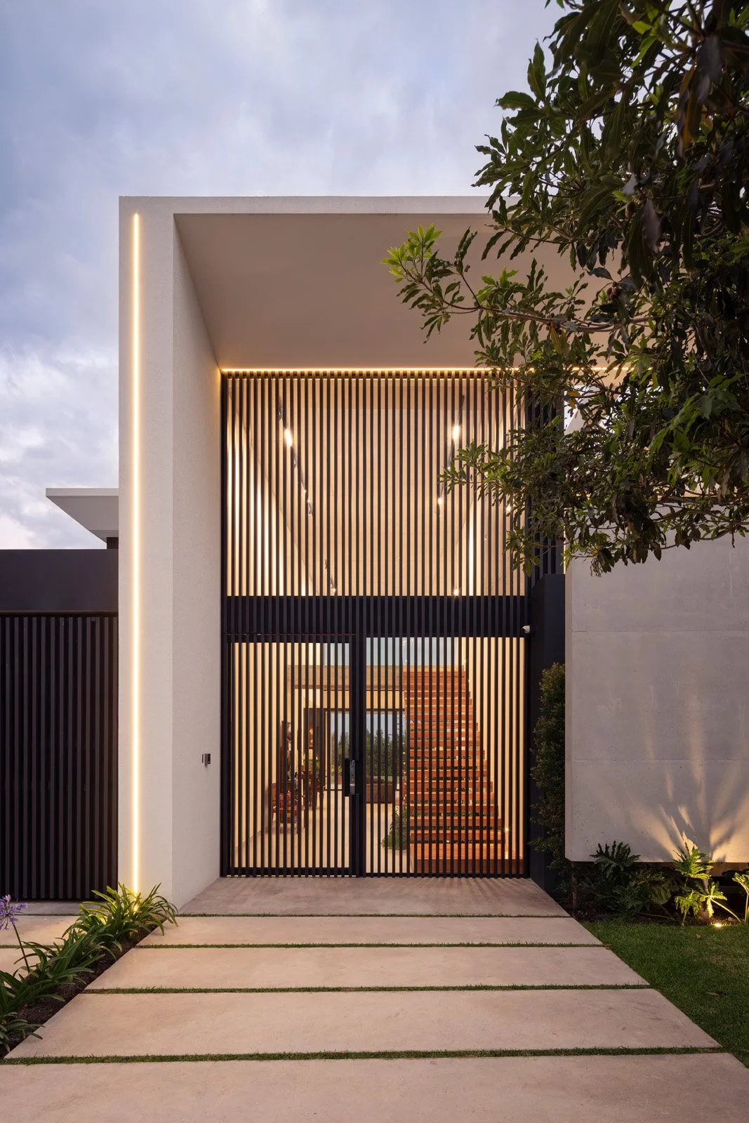 Entrada moderna de casa con puertas de barrotes negros y iluminación cálida, rodeada de plantas y un camino de baldosas.