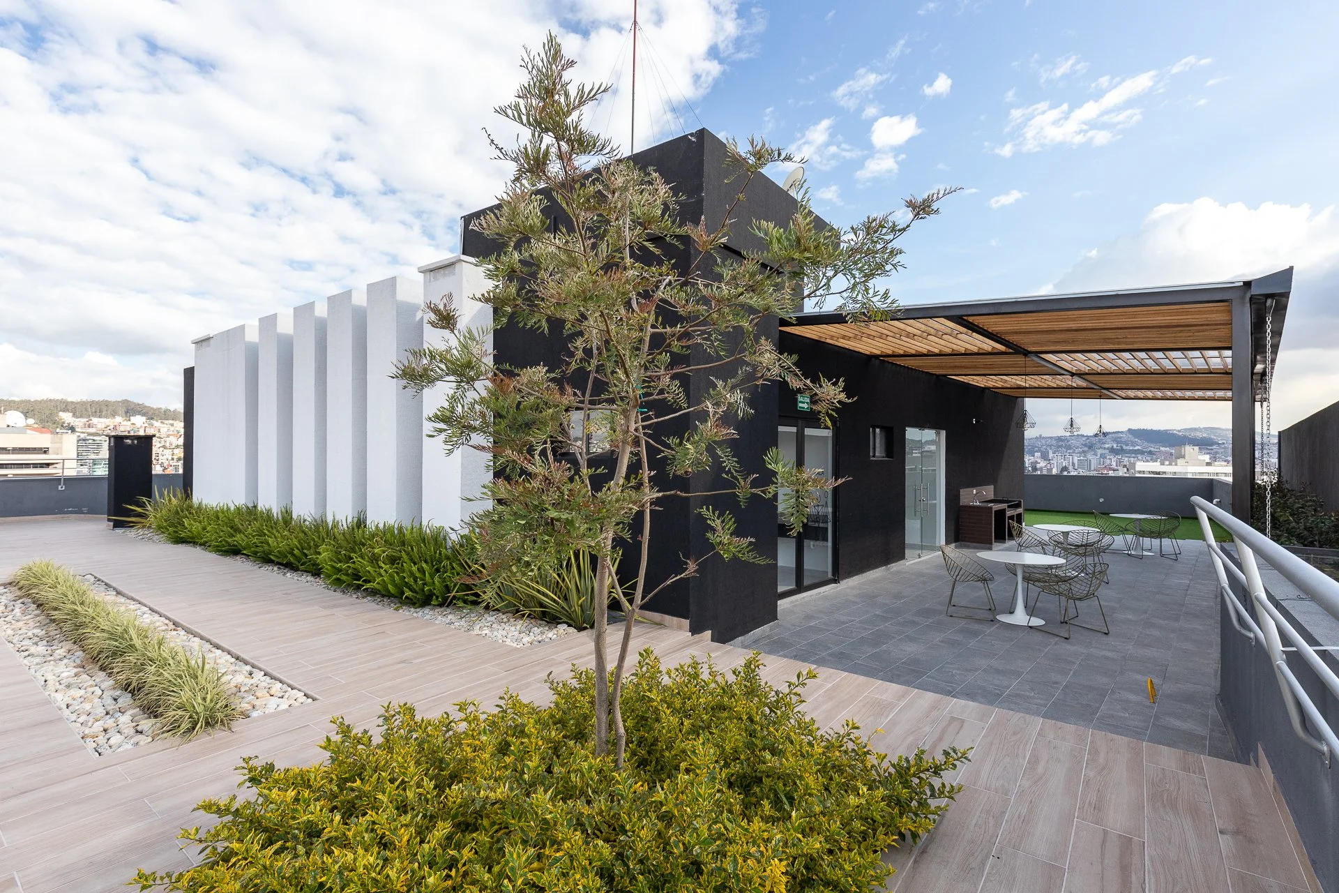 Terraza moderna con áreas verdes, mesas y sillas, estructura de madera y vista urbana al fondo