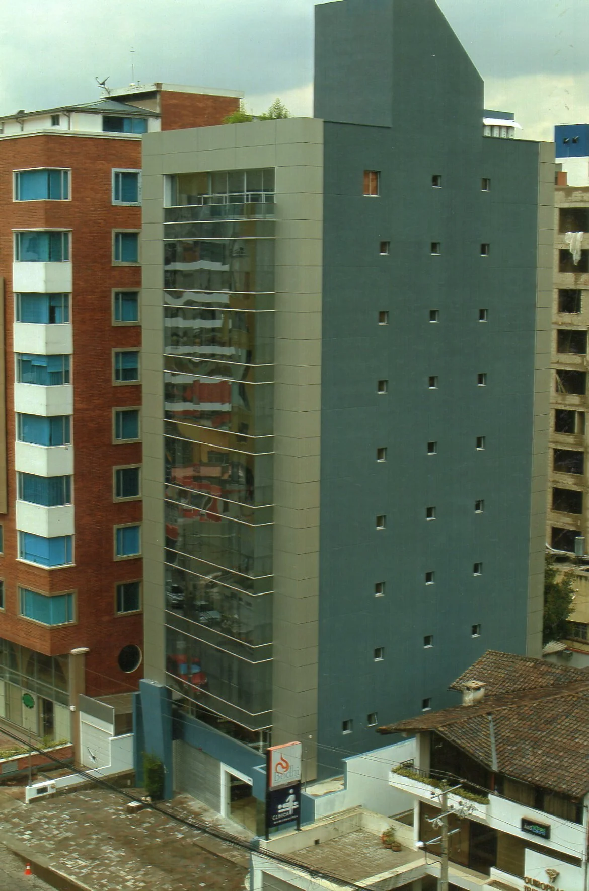 Edificio moderno con fachada de vidrio y paredes de color gris, rodeado de otros edificios residenciales en un área urbana.