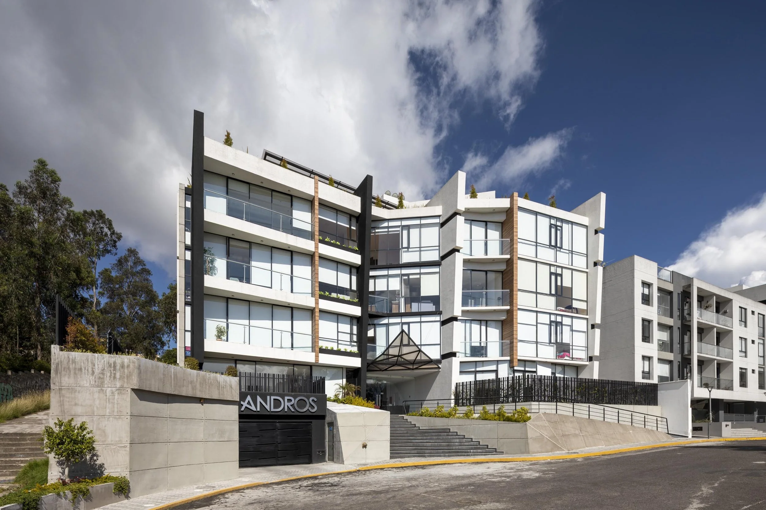 Edificio moderno de varias plantas con amplios ventanales y una entrada con escaleras, rodeado de aceras y vegetación, bajo un cielo parcialmente nublado.
