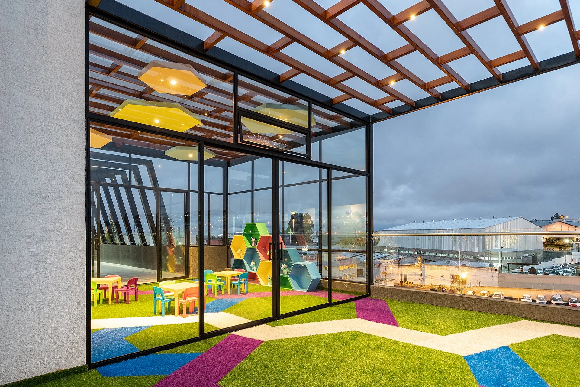 Espacio infantil con mesas y sillas de colores, estanterías hexagonales, paredes de vidrio, y un techo de madera con lámparas, vista de ciudad en la noche