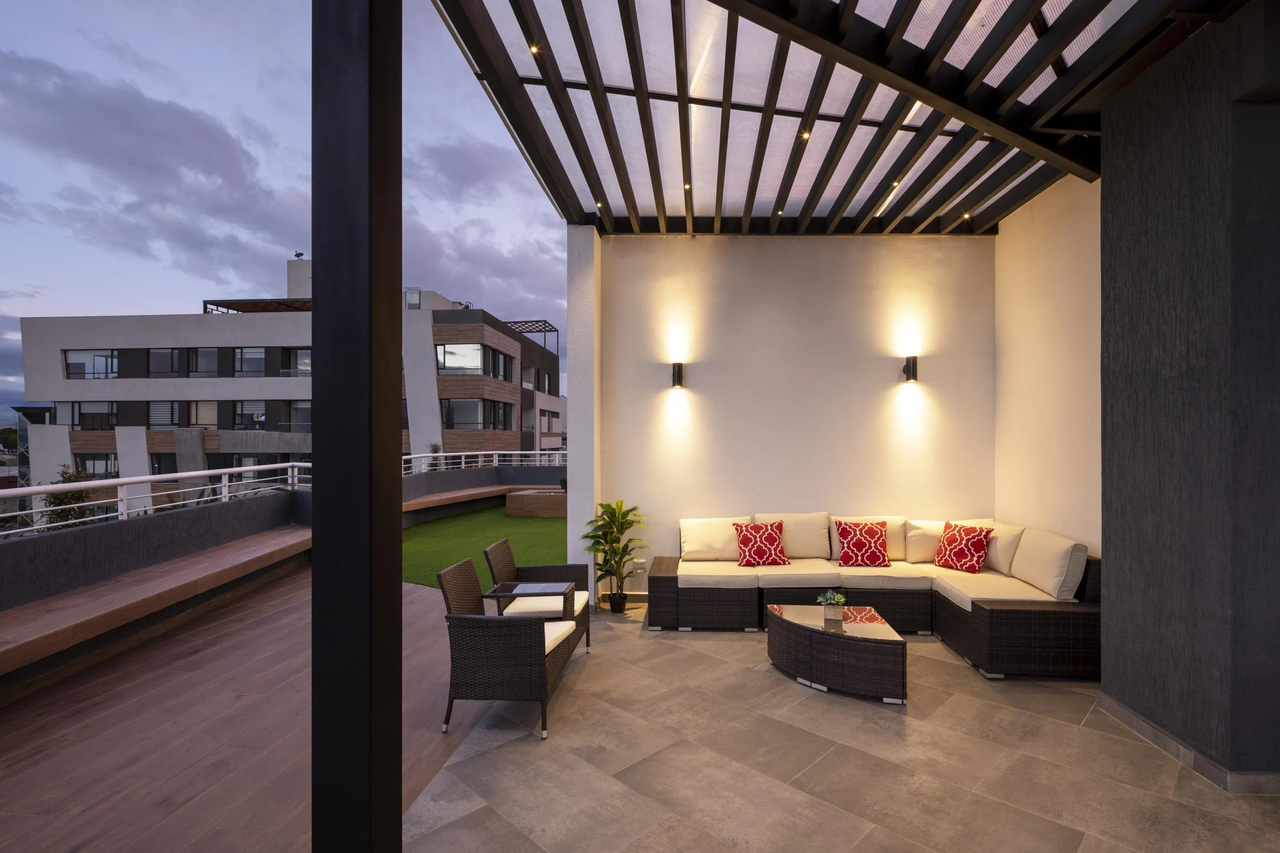 Terraza moderna con sillones beige y cojines rojos, plantas verdes, paredes blancas, lamparas de pared y techo de madera, vista de edificio de apartamentos en la tarde