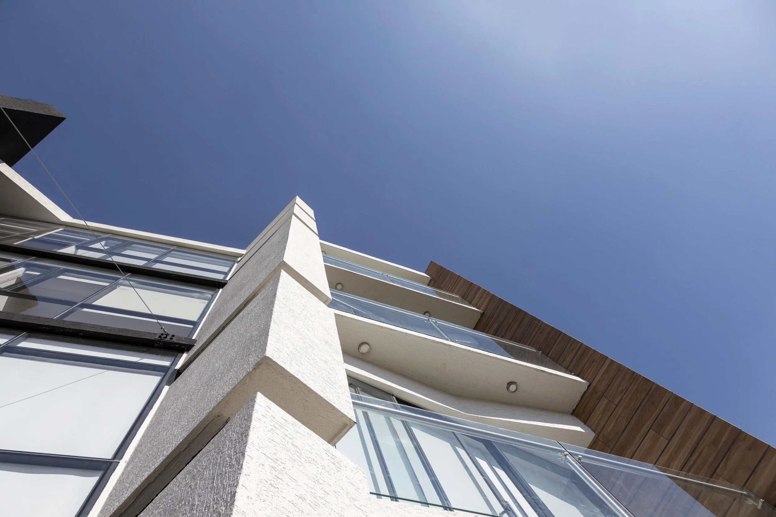Vista hacia arriba de un edificio moderno con balcones de vidrio y detalles de madera, bajo un cielo despejado y azul.