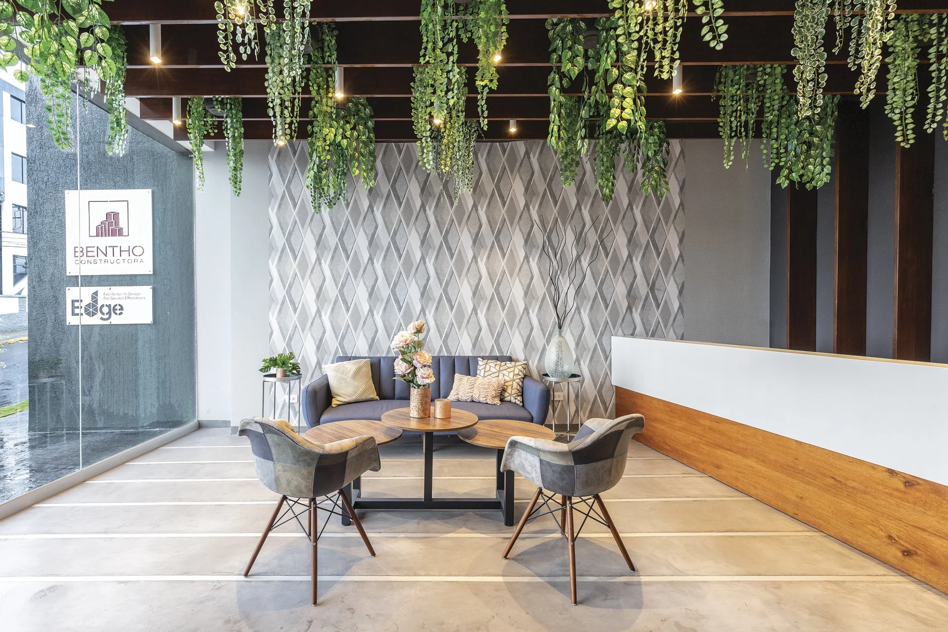 Vestidor con sillones y una mesa de centro, paredes decoradas con patrones geométricos, plantas colgantes en el techo, y una gran ventana de vidrio.