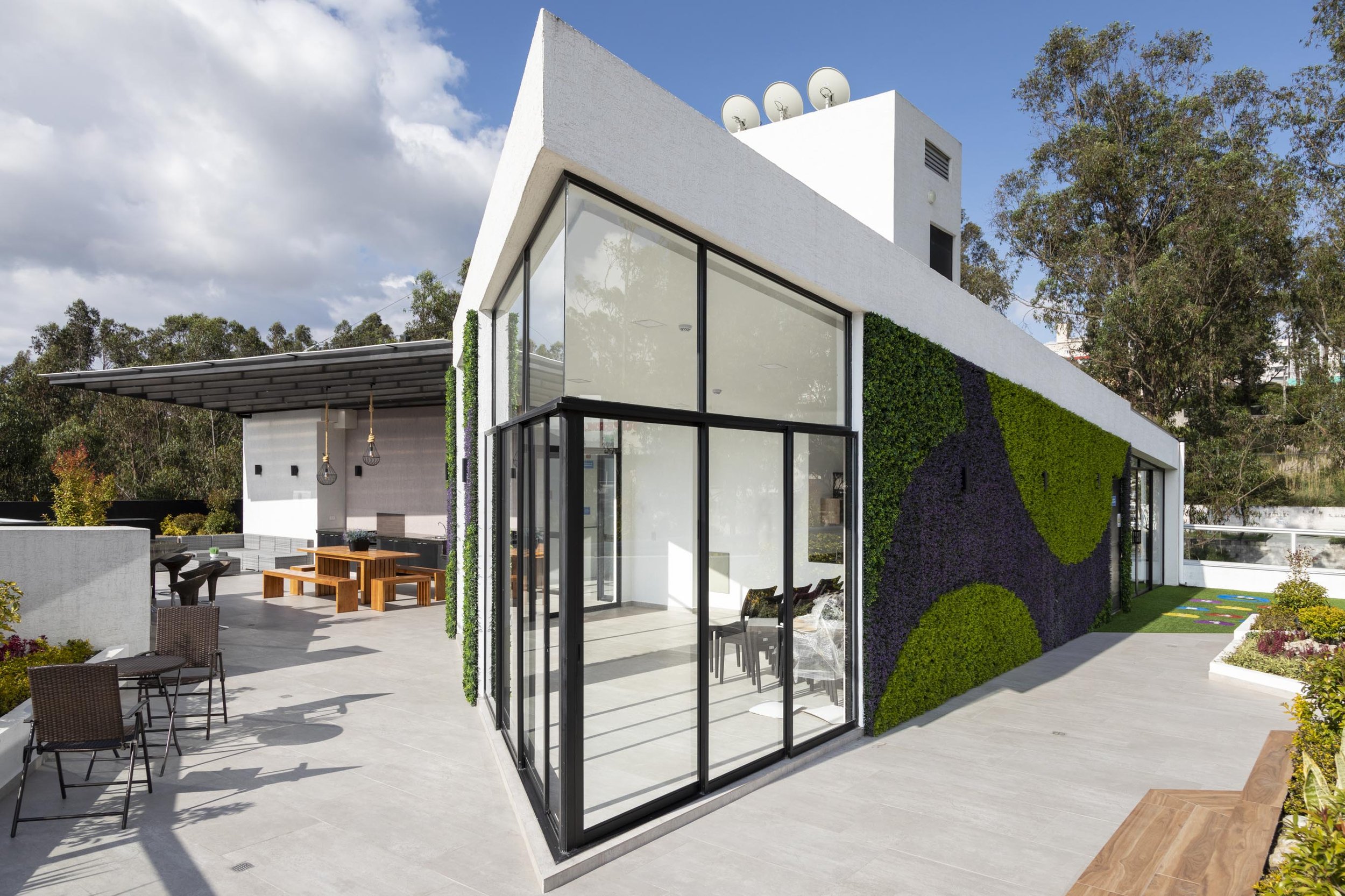 Imagen de una casa moderna con paredes blancas y grandes ventanas de vidrio, decorada con un muro verde y plantas, con área de descanso exterior y un cielo parcialmente nublado.