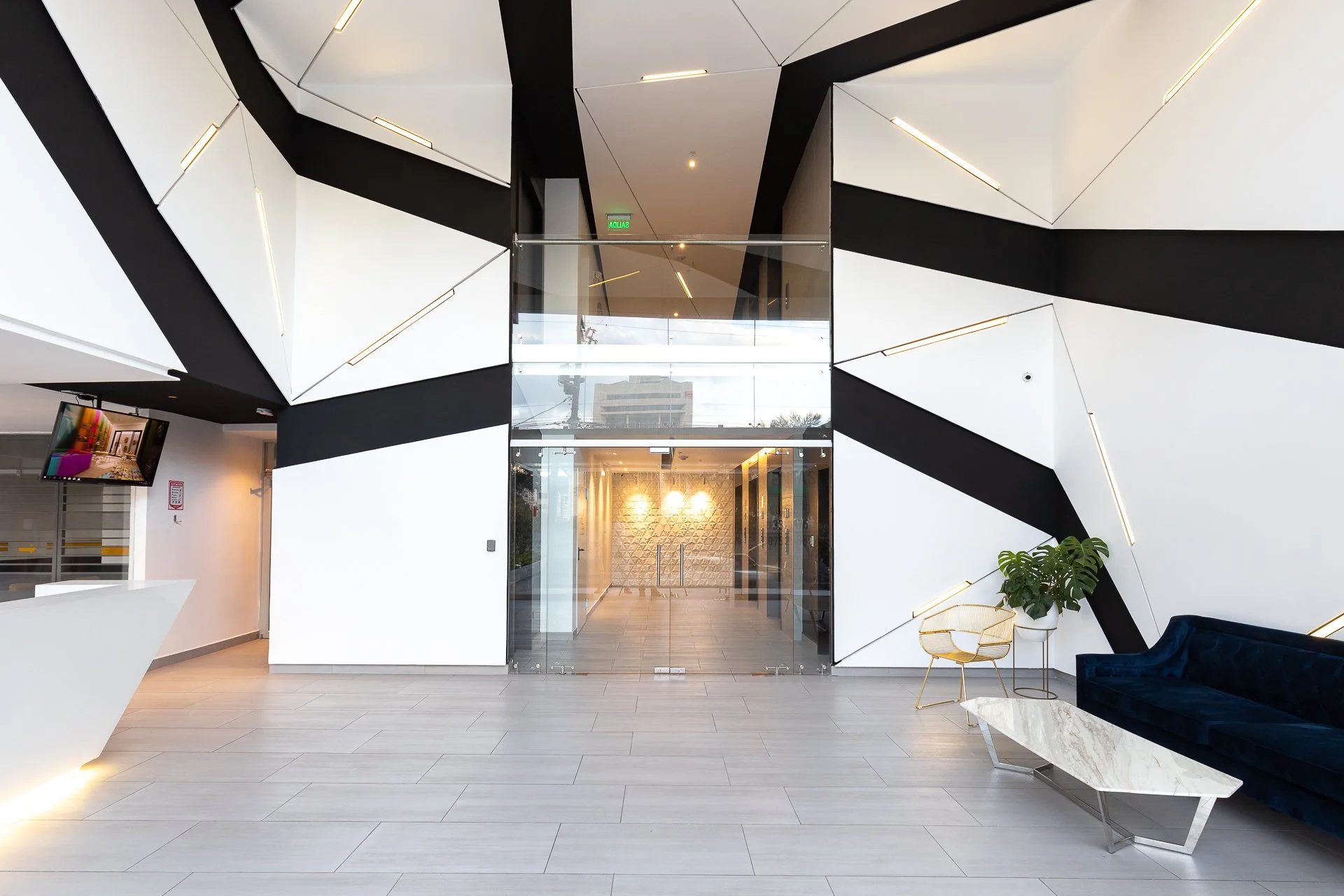 Lobby moderna con paredes decoradas en blanco y negro, sillón azul, mesa de mármol, planta y televisión.