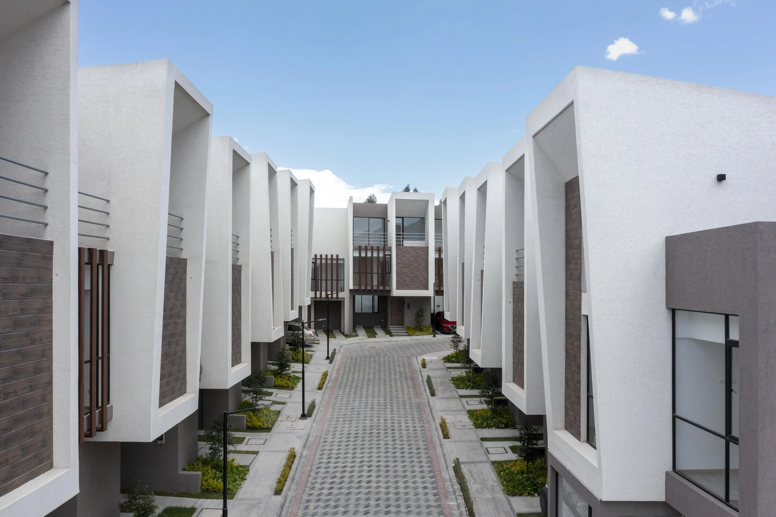 Vista de un complejo de viviendas modernas con fachada blanca, ventanas grandes y un camino pavimentado en el centro, rodeado de áreas verdes y farolas.
