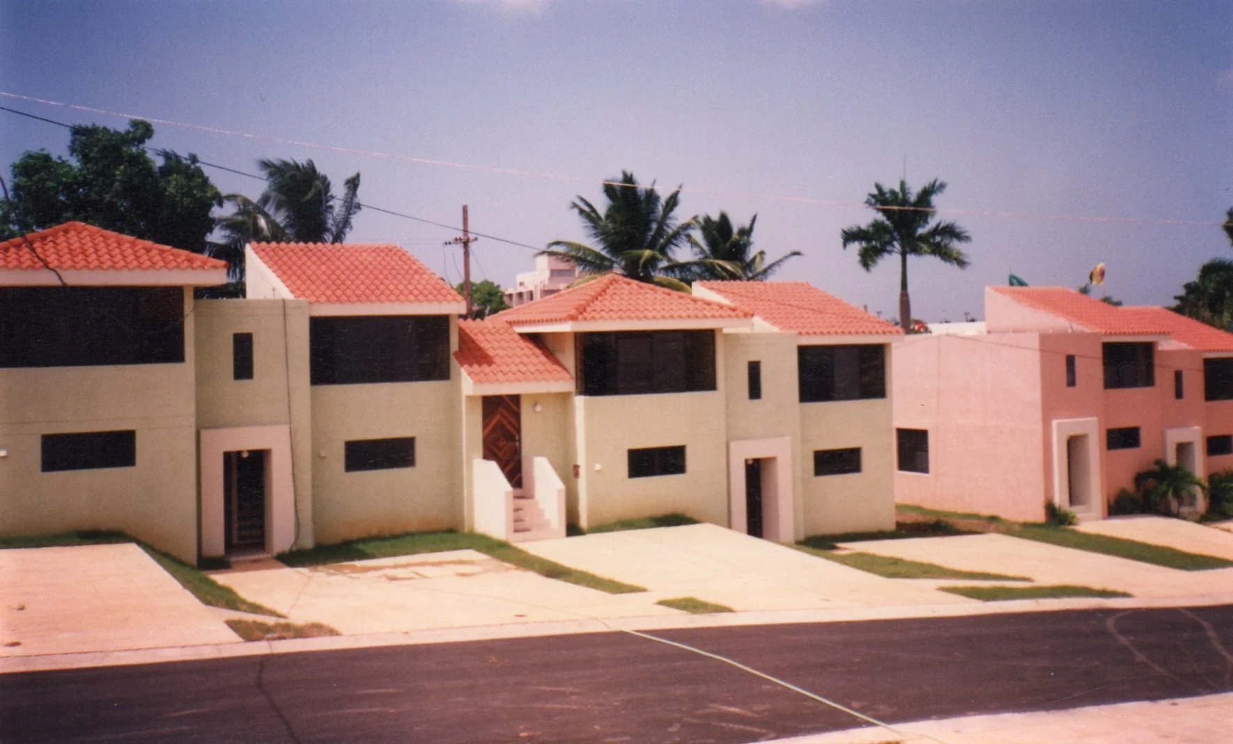Conjunto de casas residenciales con techos de teja roja, paredes de colores pastel, en un entorno con palmeras y un cielo despejado.