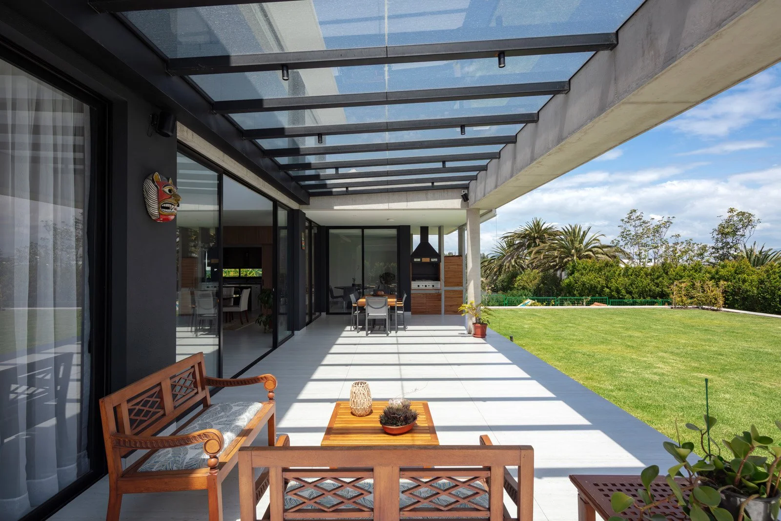Terraza moderna con mobili wooden y área de parrilla, vista a un jardín con árboles y césped en un día soleado.