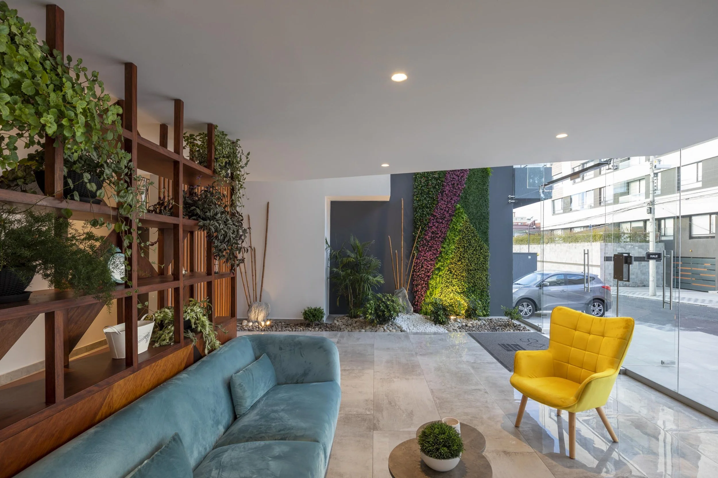 Salón moderno con sillón amarillo y sofá azul, pared con jardín vertical, estantería de madera con plantas, y vista a la calle a través de ventanas de cristal.