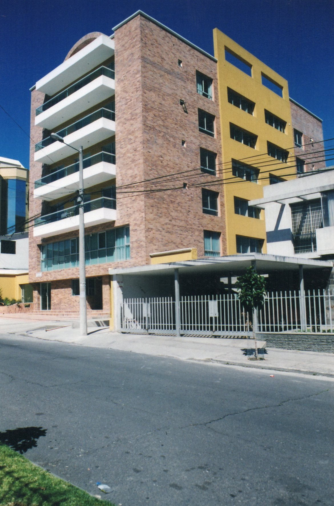 Edificio moderno de varios pisos con balcones blancos, fachada de ladrillo y detalles en amarillo, en una calle con agua y árboles.