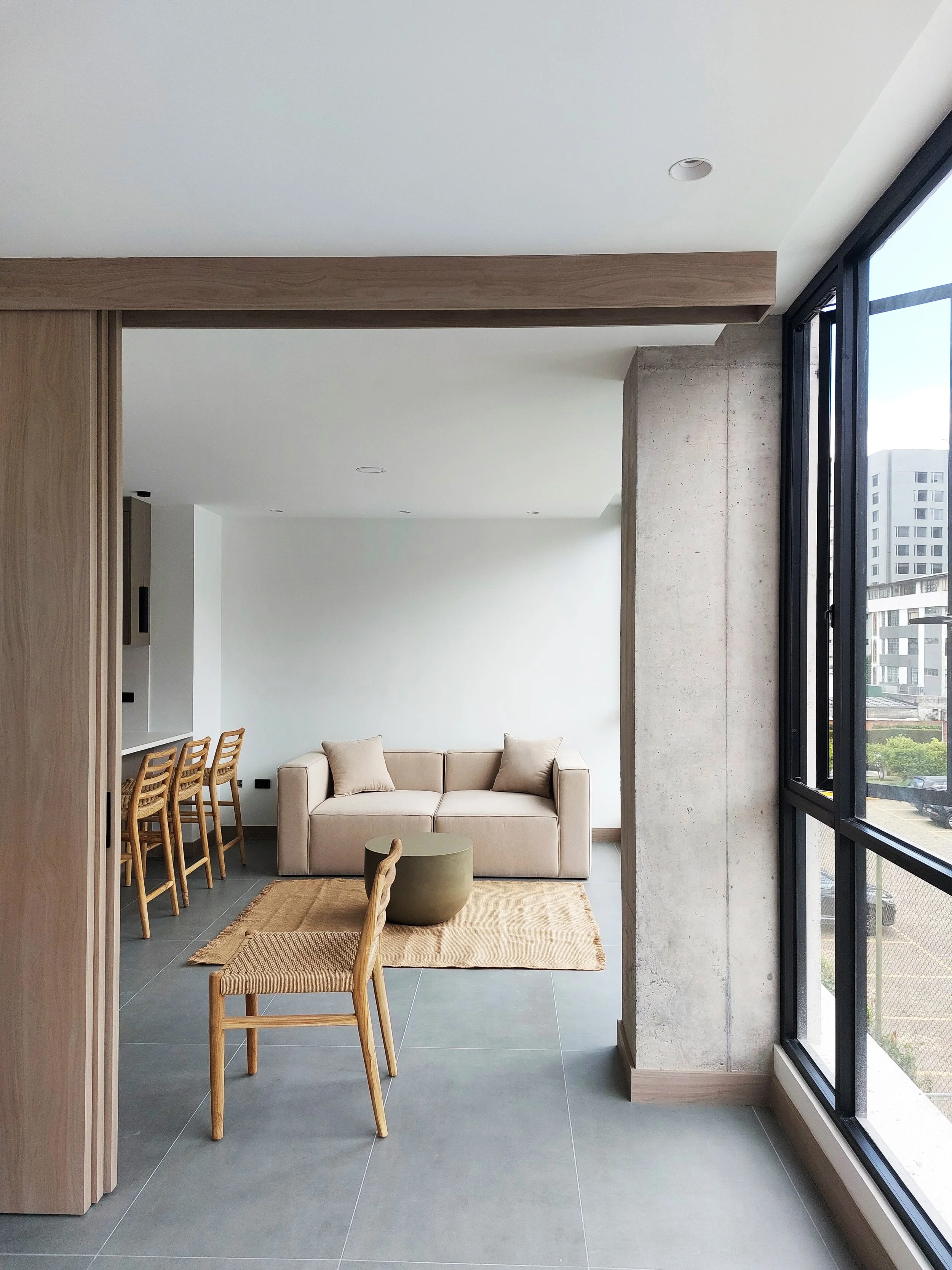 Sala moderna con sofá beige, sillones de madera, mesa decorativa y ventanas grandes que dejan entrar mucha luz natural