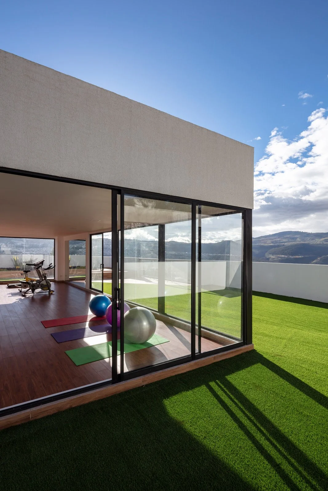 Interior de una casa moderna con vista a un patio con césped, instrumentos de ejercicio y pelotas de diferentes colores. Tiene paredes de vidrio que permiten vista panorámica de las montañas y el cielo nublado.