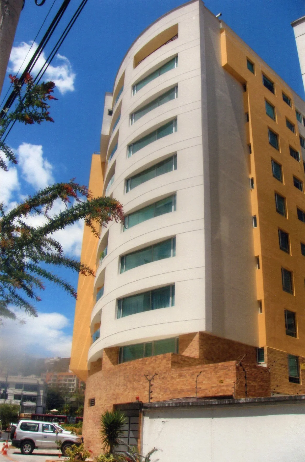 Edificio moderno de varios pisos con ventanas grandes, paredes blancas y detalles en color dorado y ladrillo, con un cielo azul y algunas nubes.