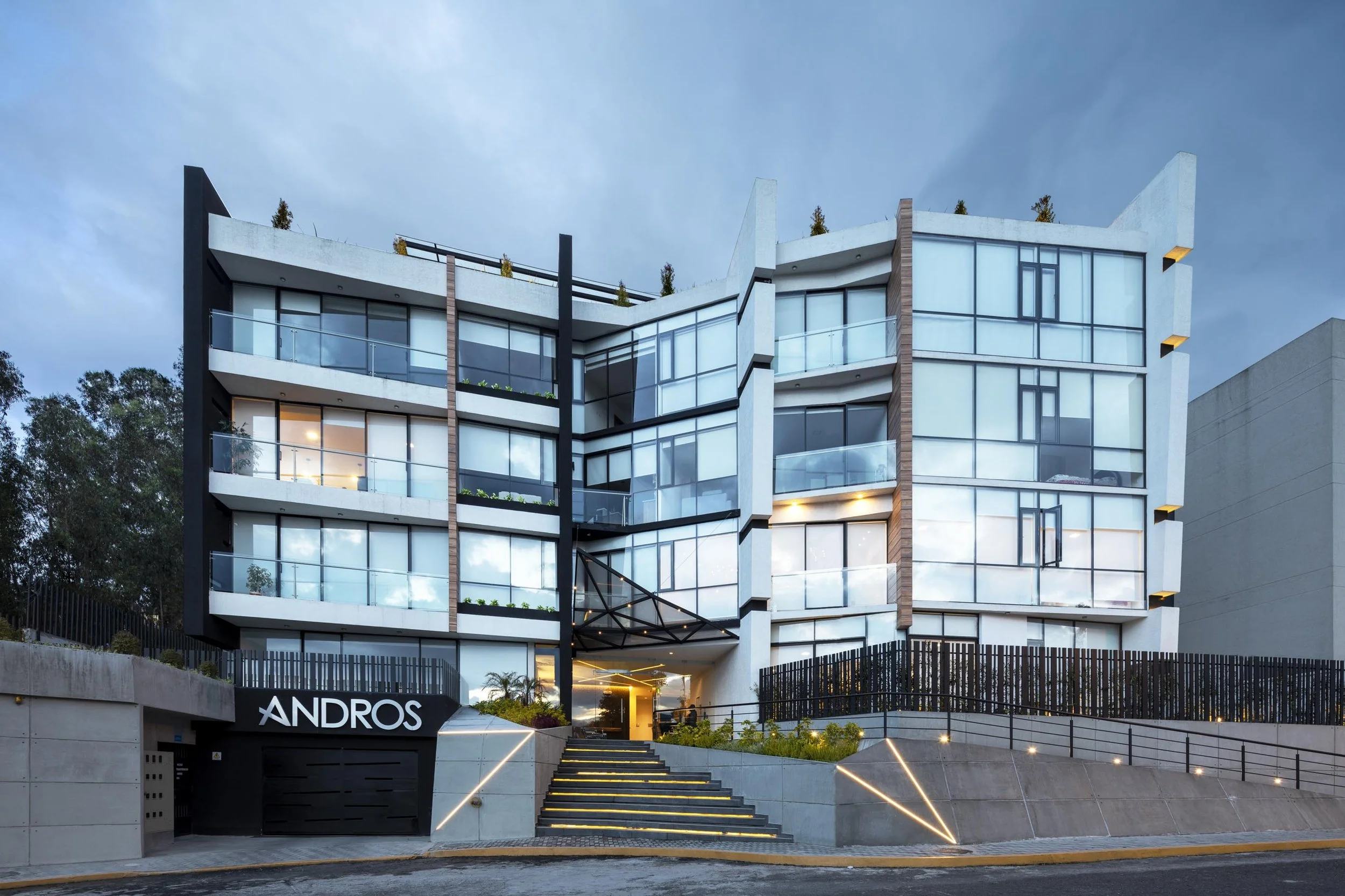 Edificio moderno de varios pisos con fachada de vidrio, entrada principal con escalones y señal que dice 'ANDROS', iluminación exterior en la noche.