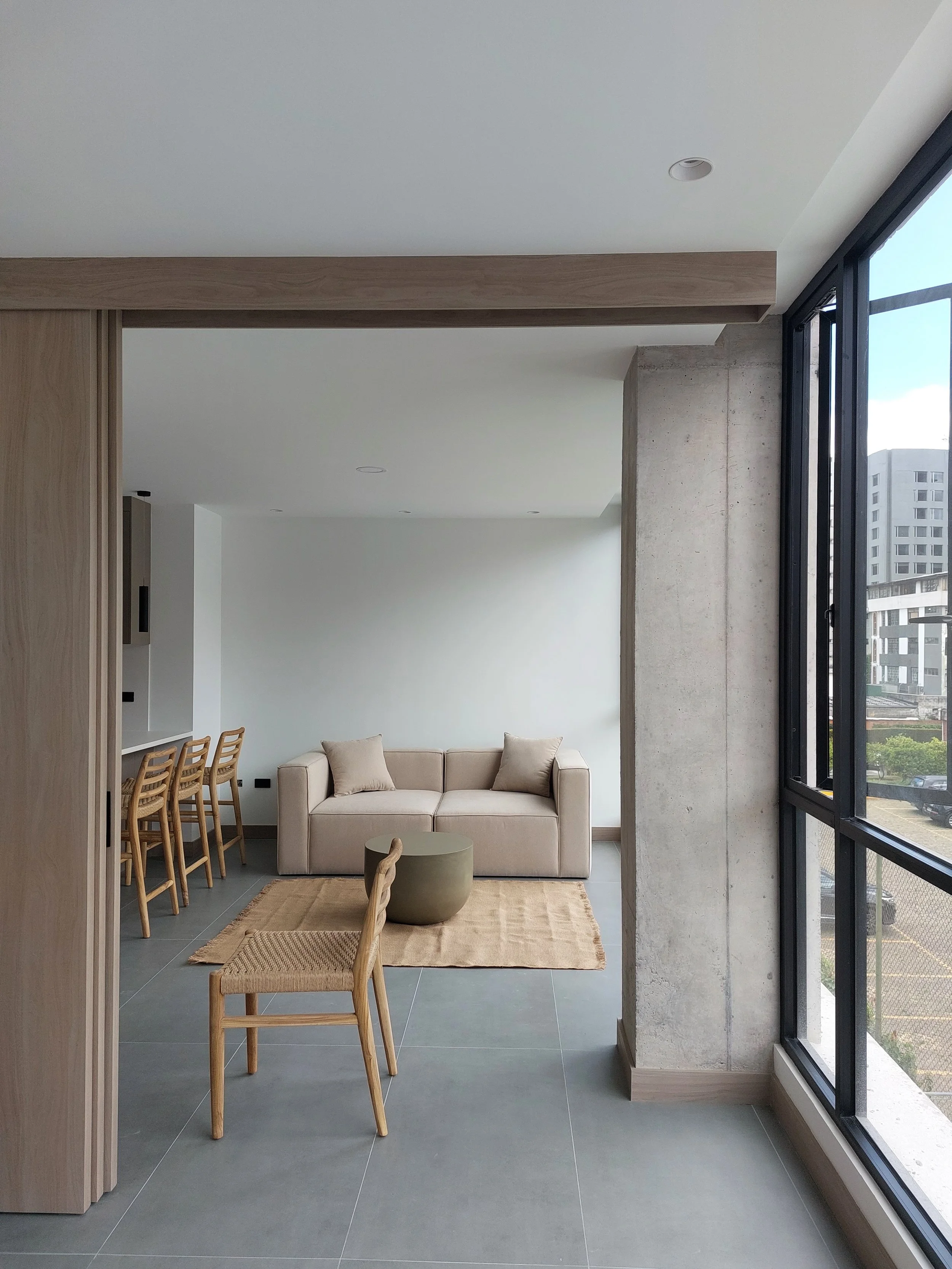 Sala moderna con sillón beige, mesa redonda, alfombra y vistas a edificios desde ventanales grandes.