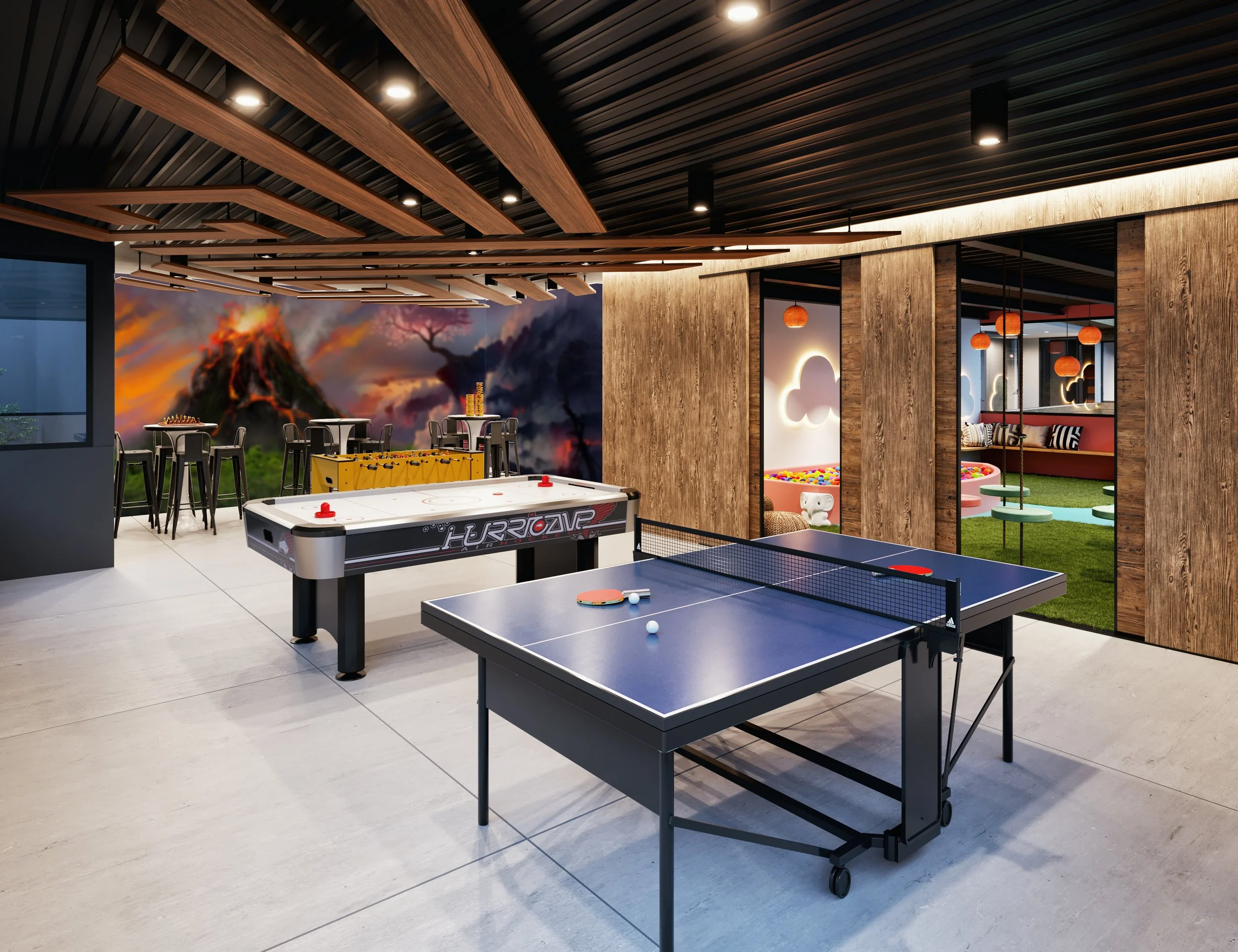 Sala de juegos con mesas de ping pong, futbolín y área infantil con columpios y bolas, decoración moderna con madera y paredes con murales de volcanes y árboles.