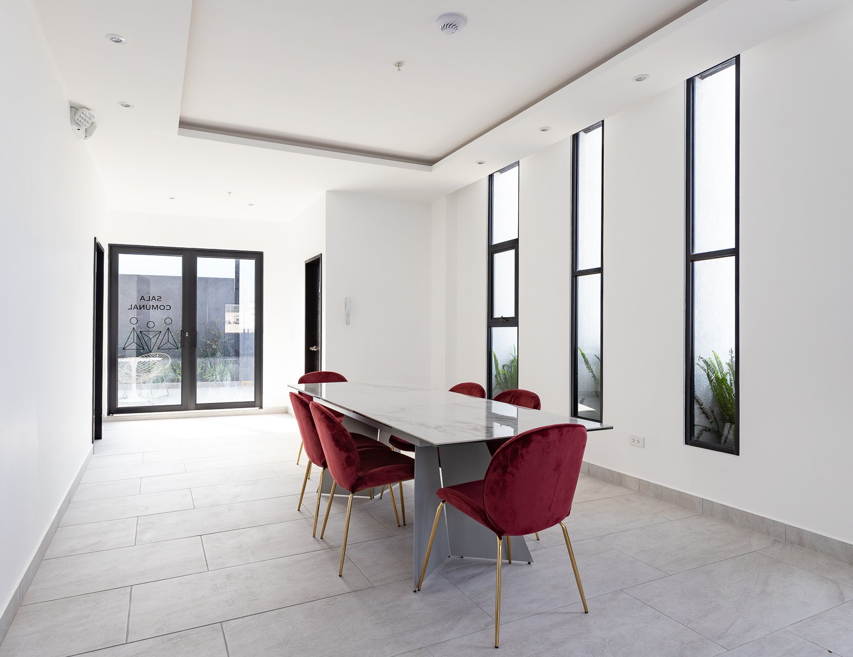 Sala de reuniones con mesa de mármol, seis sillas de terciopelo rojo, paredes blancas, ventanas altas, puerta de entrada con letrero "Sala Comercial" y plantas en la esquina.