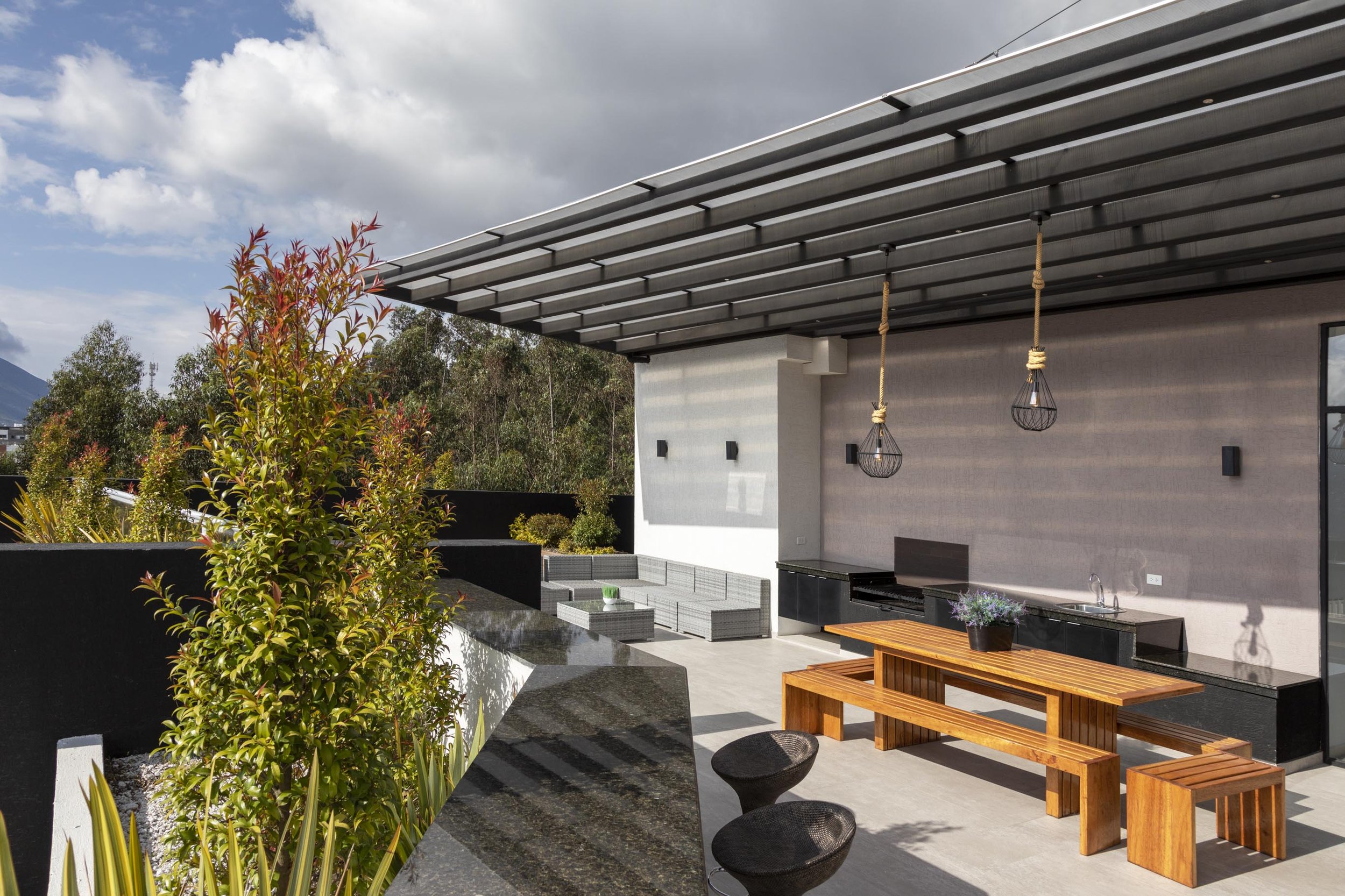 Terraza moderna con muebles de madera, plantas y área de cocina al aire libre bajo un toldo metálico, con árboles y cielo nublado al fondo.
