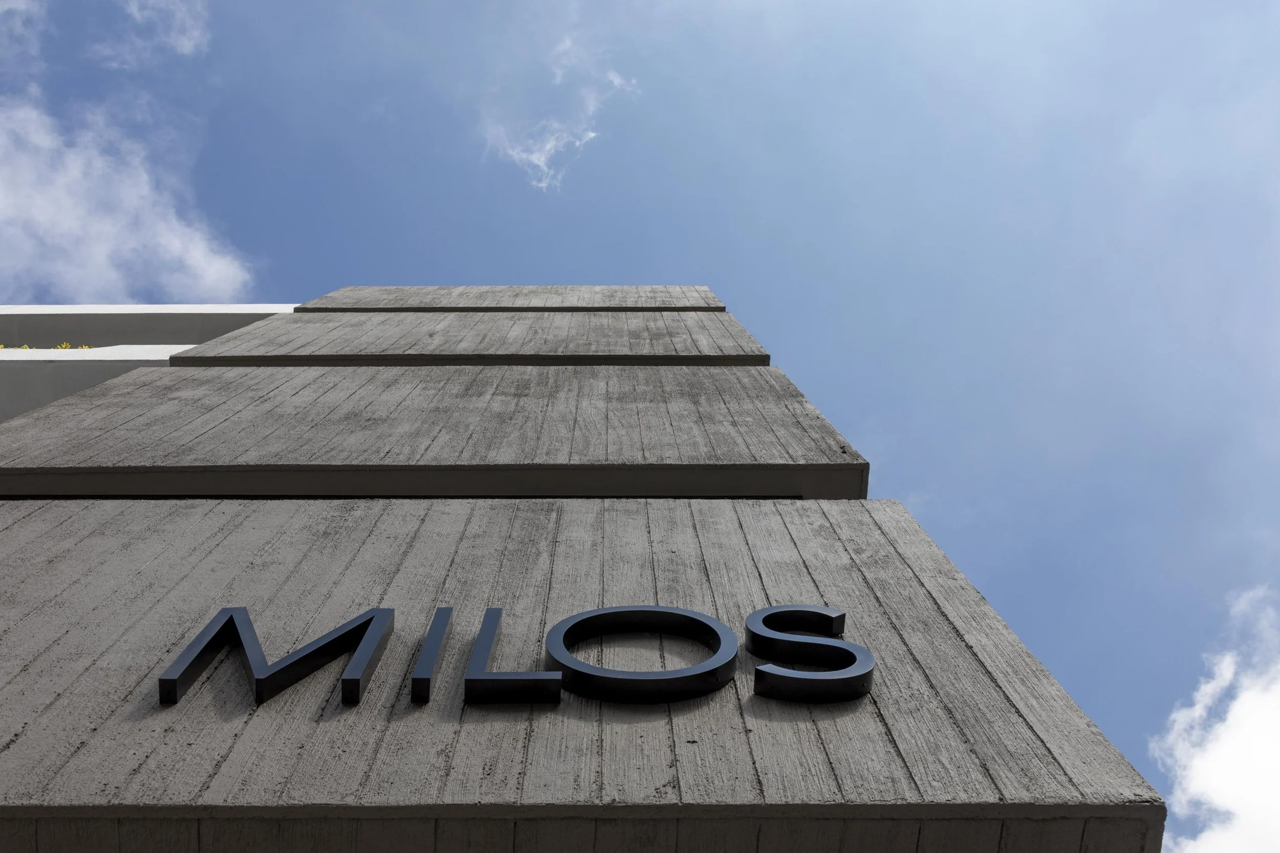 Fachada de un edificio de concreto con letras negras que dicen 'MILOS', vista desde abajo con cielo azul y nubes.