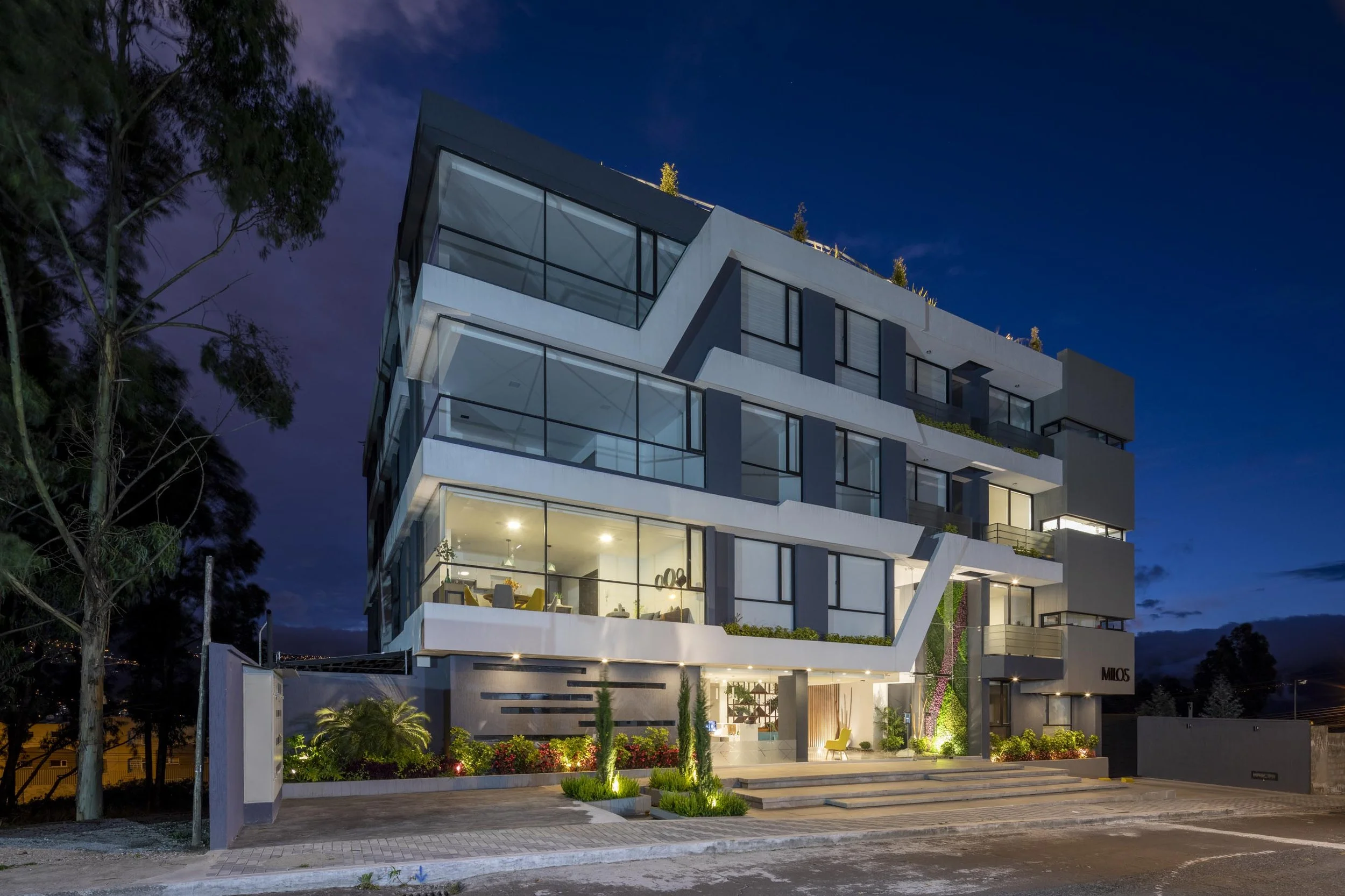 Edificio de apartamentos moderno iluminado por la noche con jardines y árboles en el área frontal.