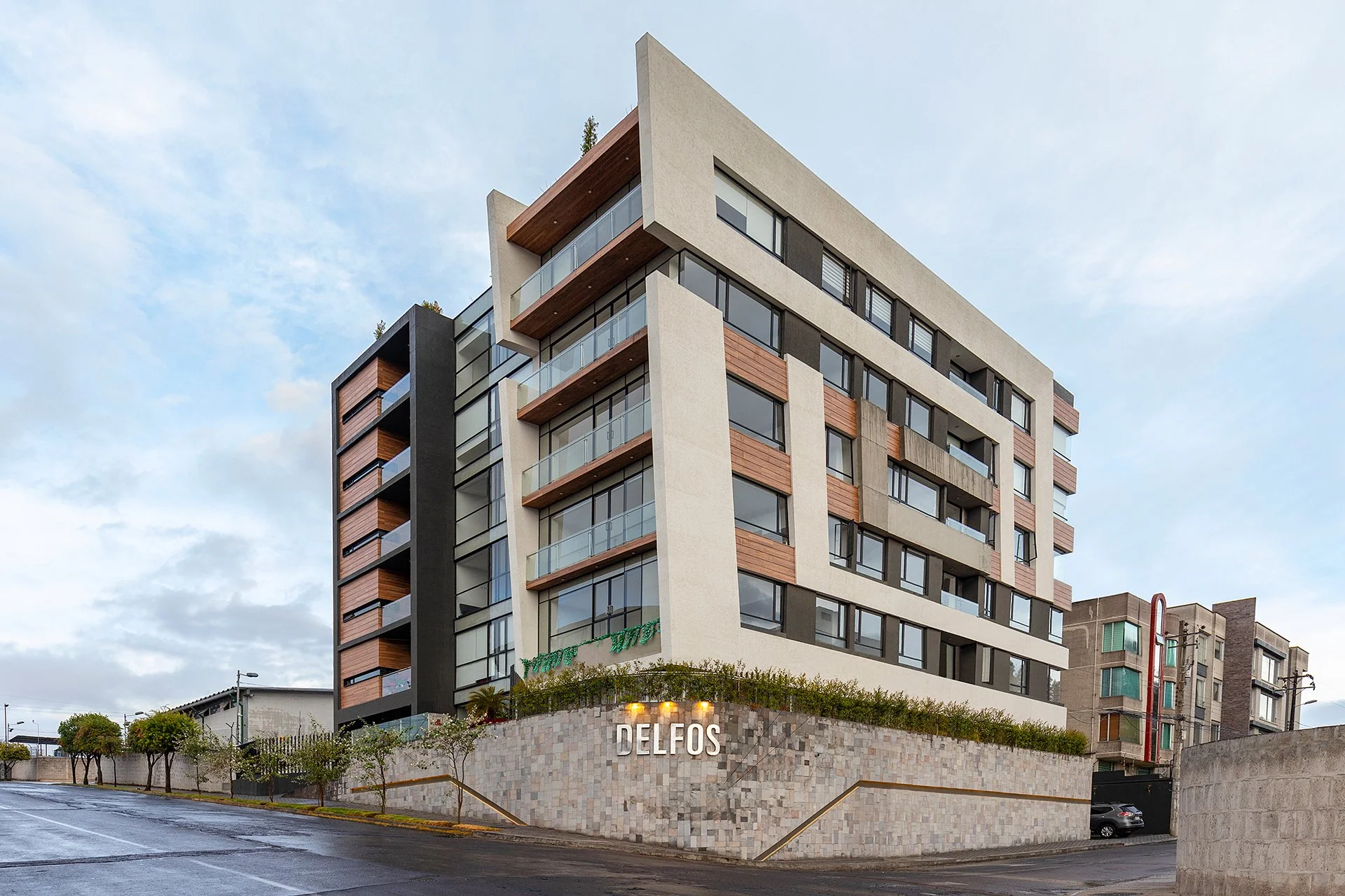Edificio moderno de varios pisos con fachada de concreto y madera, balcones de vidrio y un letrero que dice 'Delfos' en la base