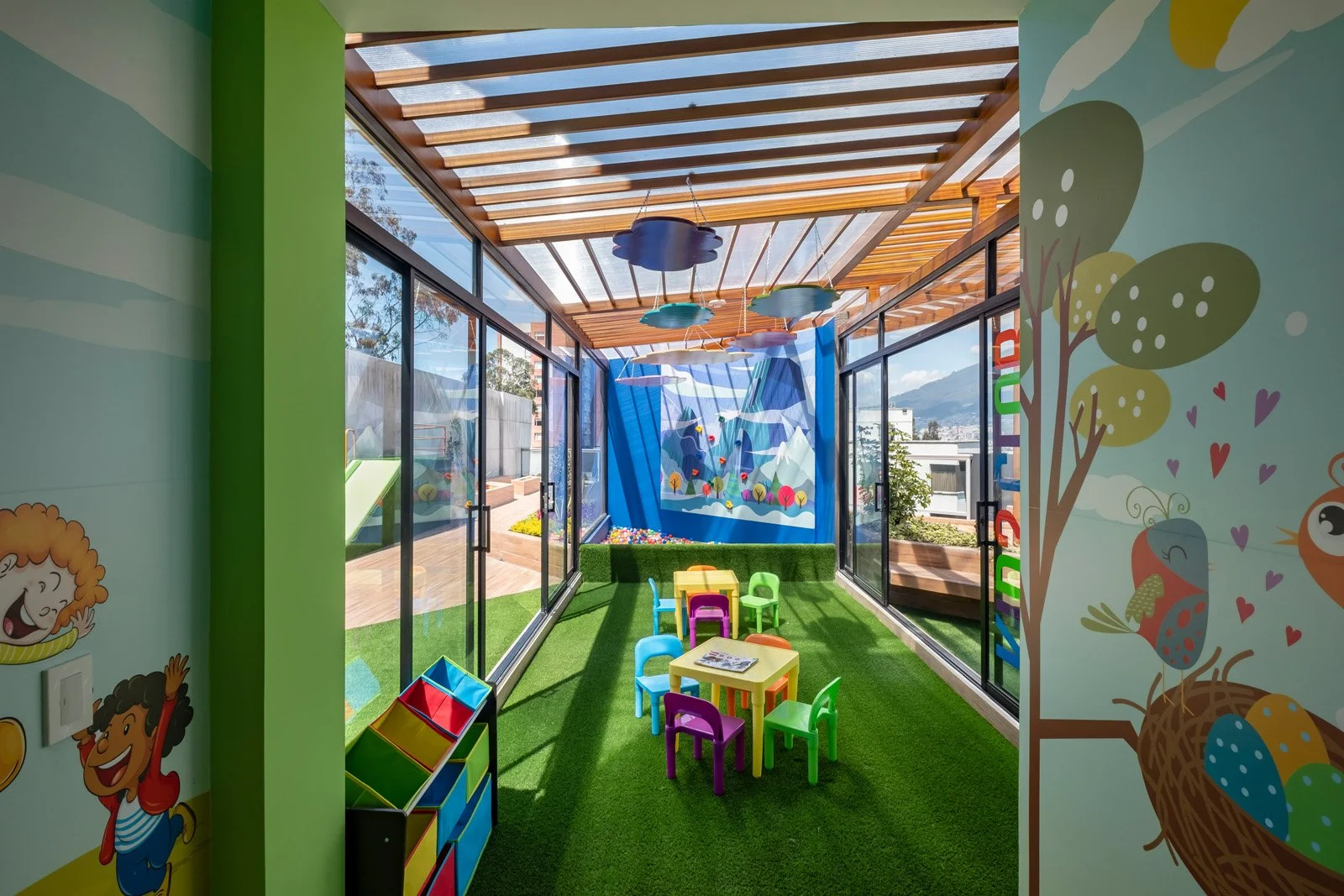 Sala infantil con paredes decoradas con dibujos de animales y árboles, suelo de césped artificial, mesas y sillas de colores, con ventanas grandes que dejan entrar luz natural y ofrecen vista a exteriores con árboles y montañas.
