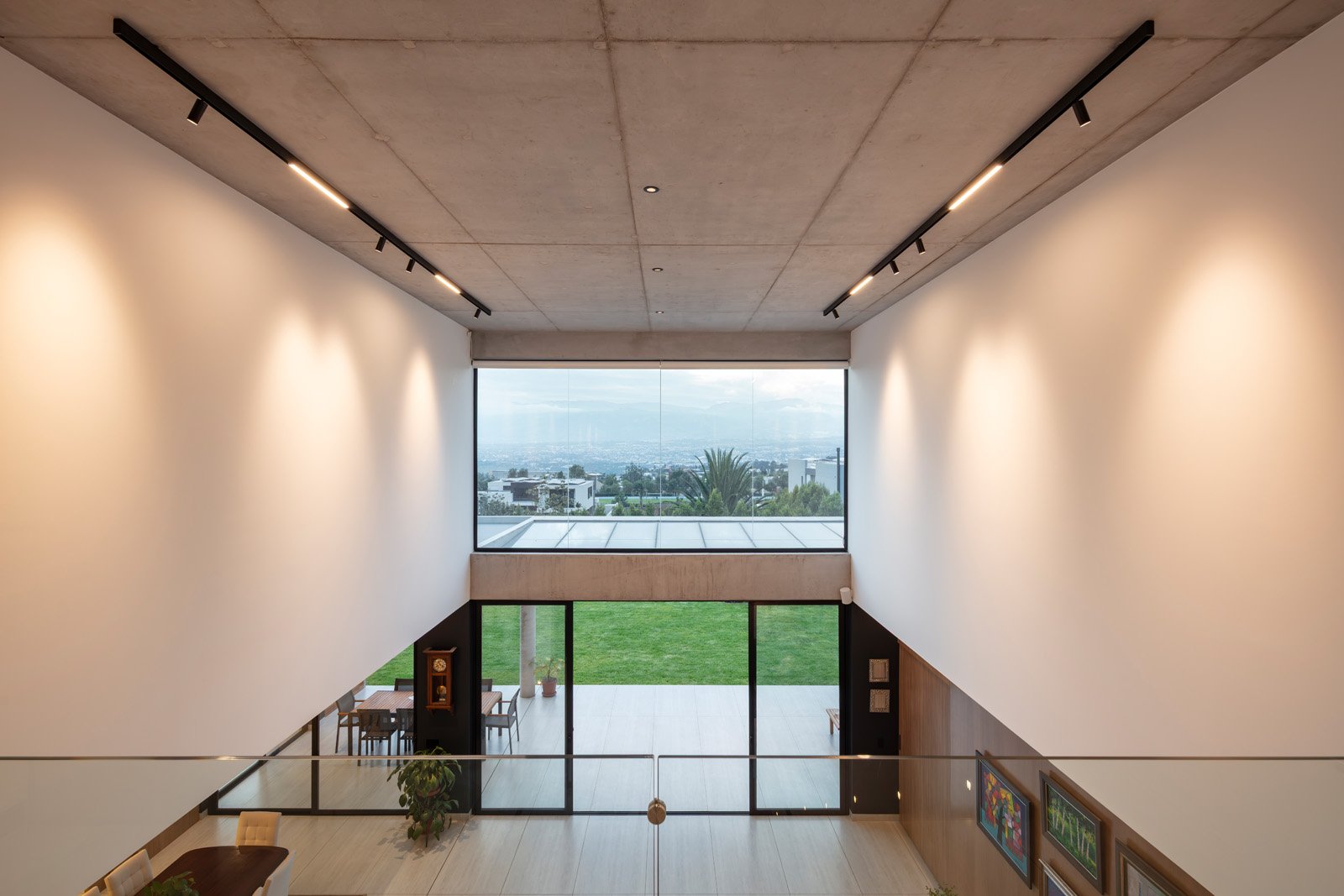 Vista interior de un espacio moderno con paredes blancas, techos de concreto y ventanas grandes con vista a un área de césped y ciudad en el fondo.