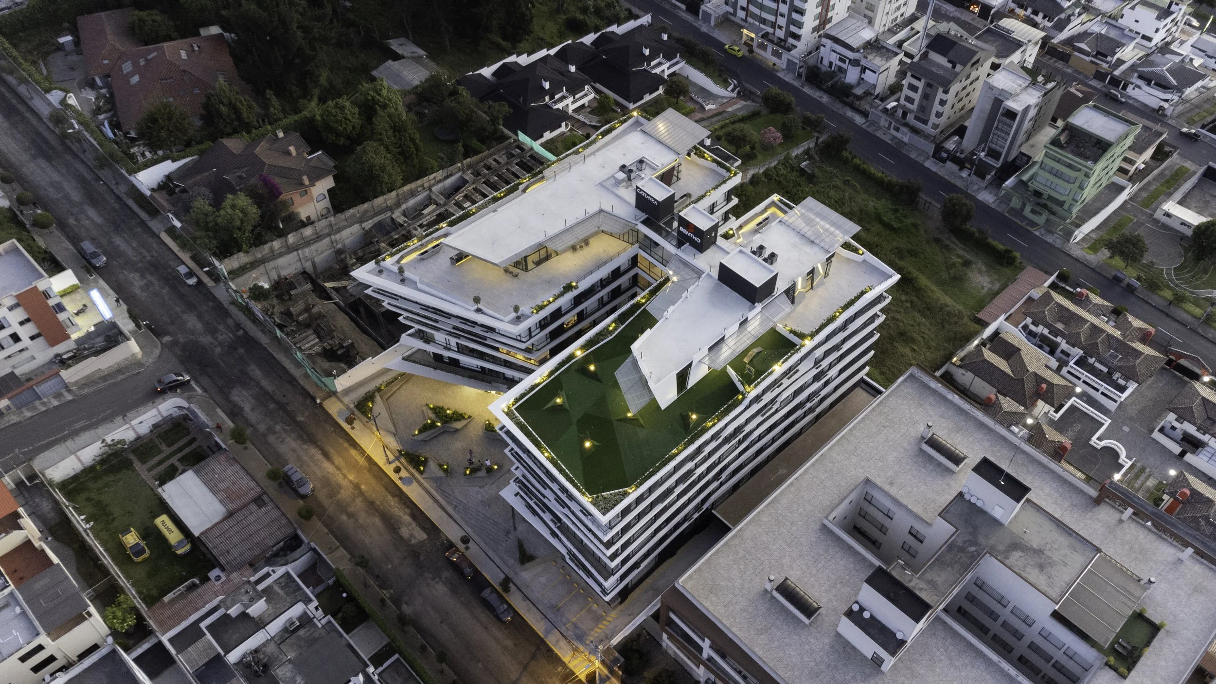 Vista aérea de un edificio moderno con áreas verdes iluminadas en la azotea, en medio de vecinas casas y edificios en una zona urbana.