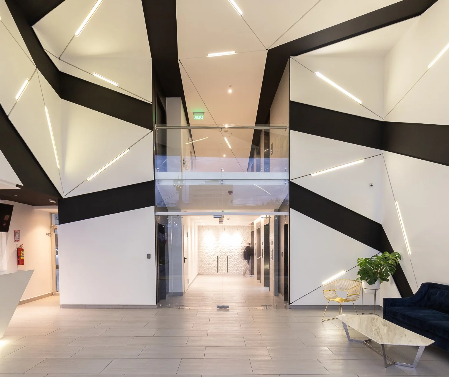 Vestíbulo de edificio moderno con paredes decorativas en blanco y negro, iluminación integrada, puertas de ascensor y área de descanso con sofá azul, silla dorada y mesa de mármol.