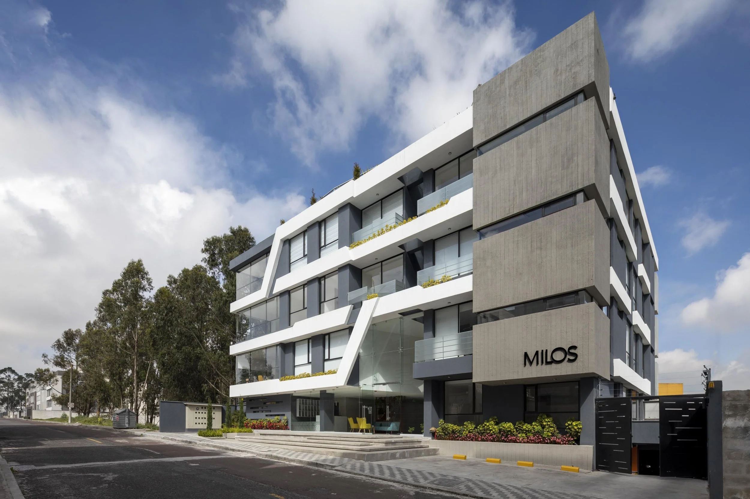 Edificio moderno con fachada de concreto y vidrio en un vecindario con árboles y cielo con nubes.