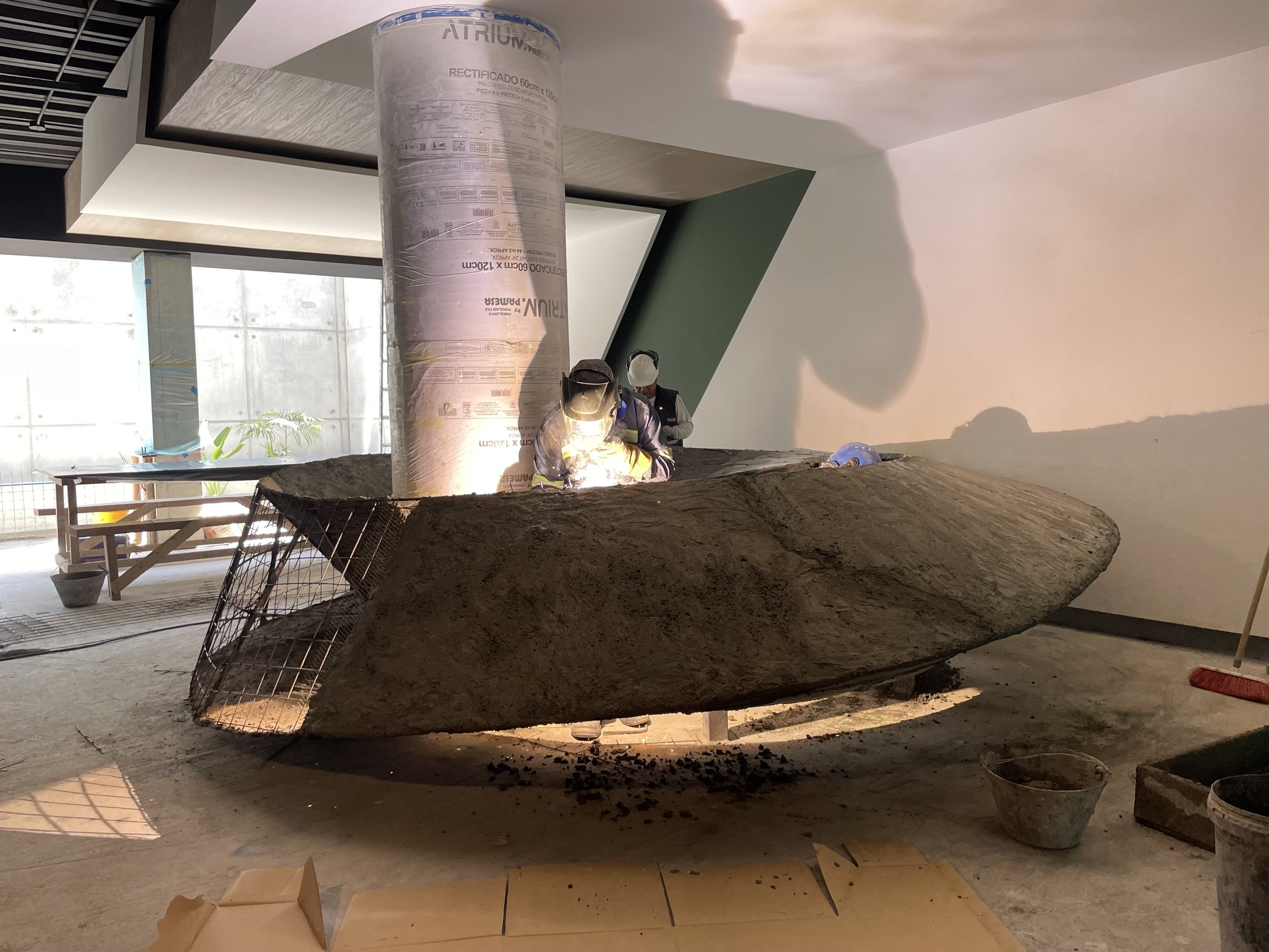 Dos trabajadores en un sitio de construcción, uno con casco y otro con mascarilla, trabajan en una estructura grande de piedra o roca, con herramientas y materiales de construcción alrededor.