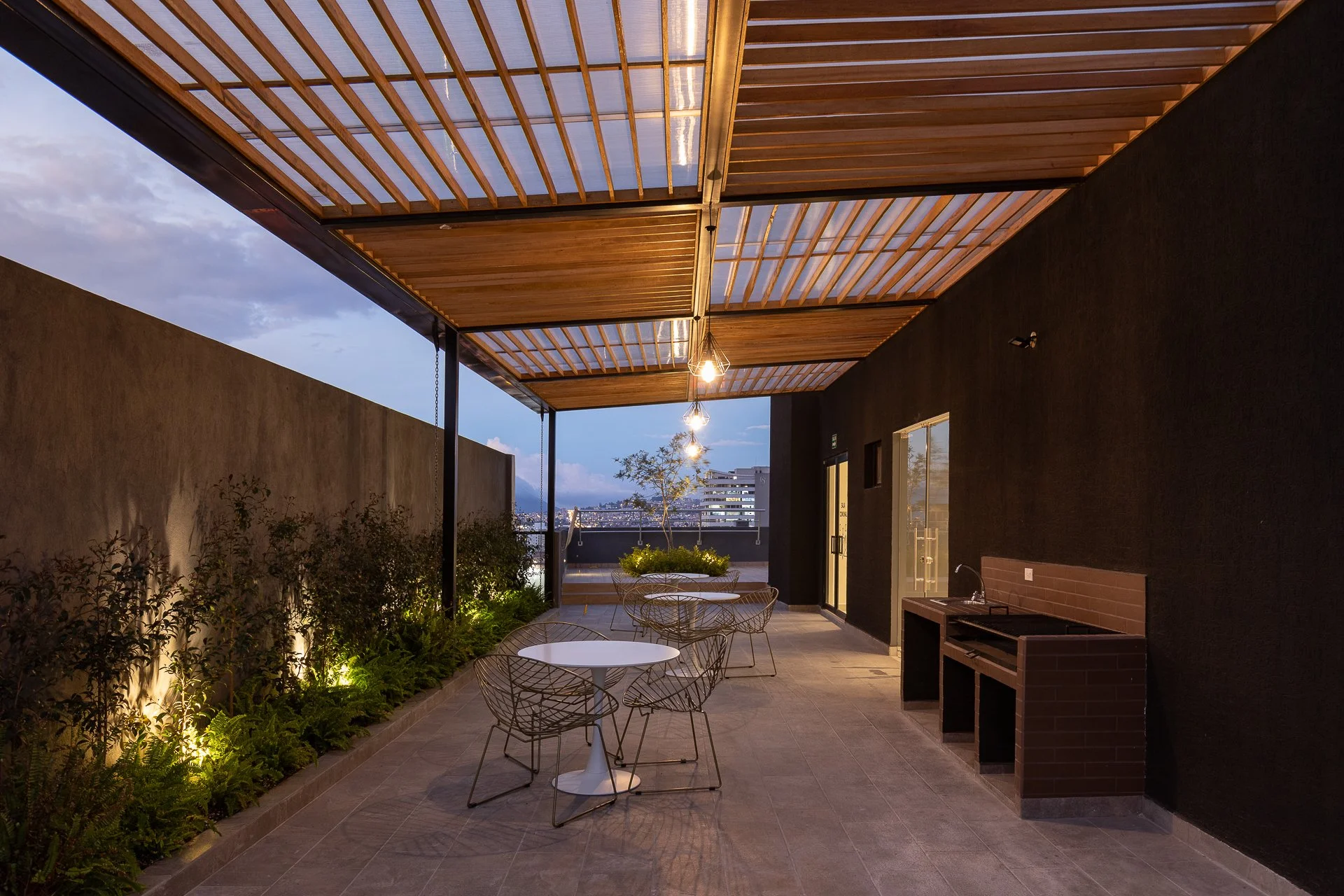 Terraza urbana con mesas blancas y sillas de cables de metal, paredes negras, iluminación suspendida y plantas decorativas.