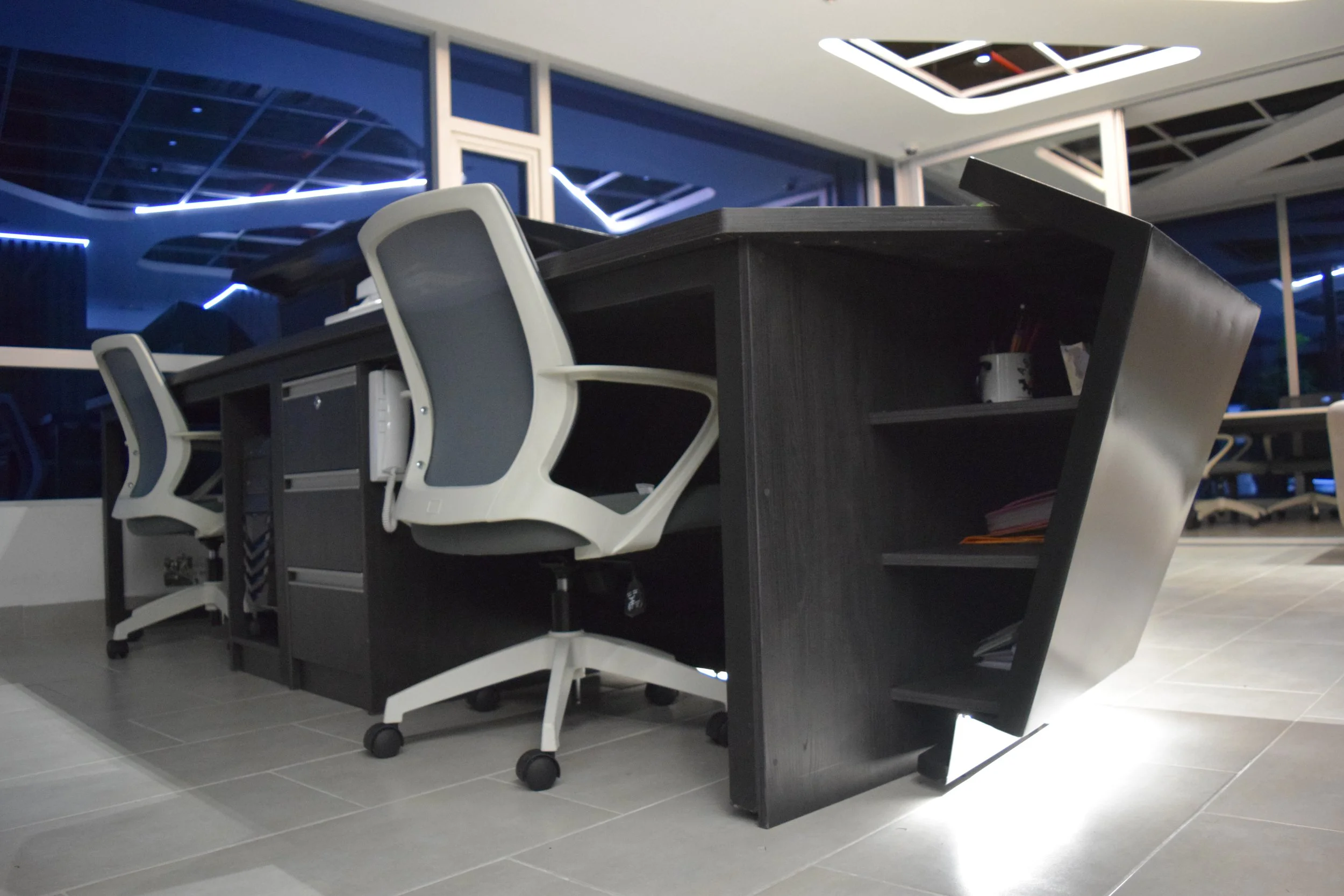 Mesas de oficina con sillas en un espacio con ventanas grandes y luces azules en el techo