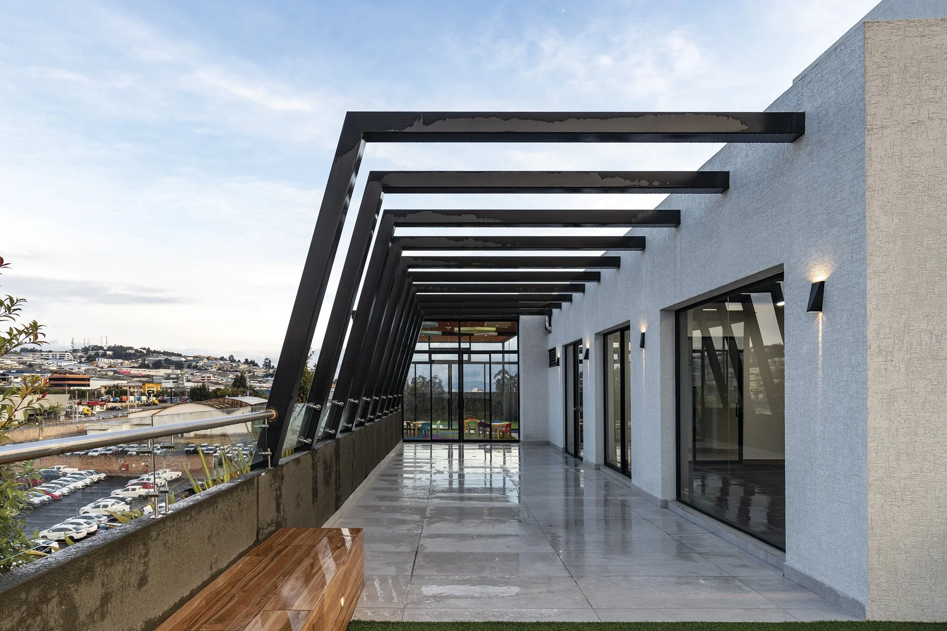 Terraza moderna con estructura de vigas negras, paredes blancas y puertas de vidrio, vista a la ciudad y cielo despejado.