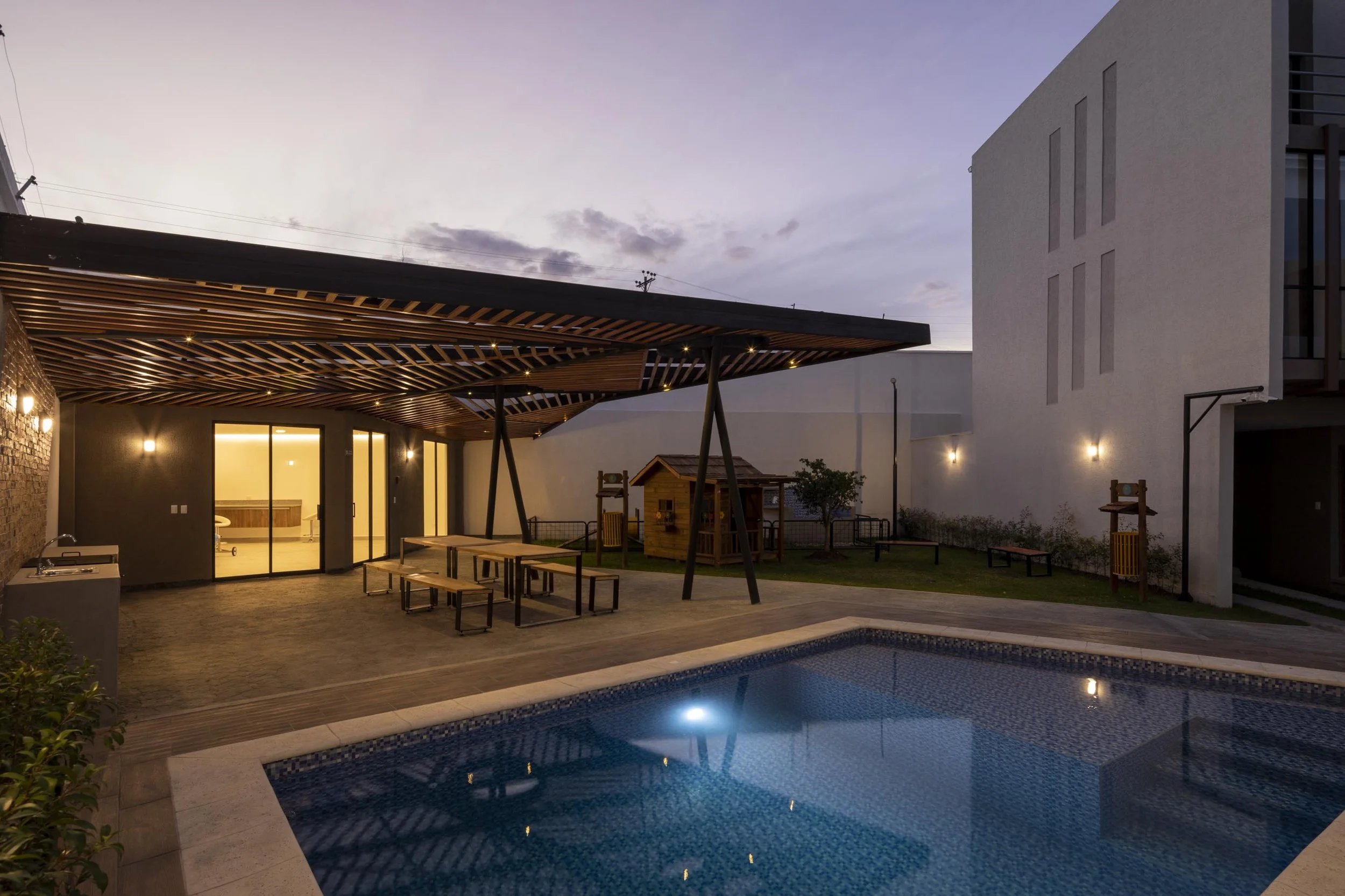 Piscina en un patio con sombra, mesas y zona de juegos infantiles, en la tarde con cielo nublado