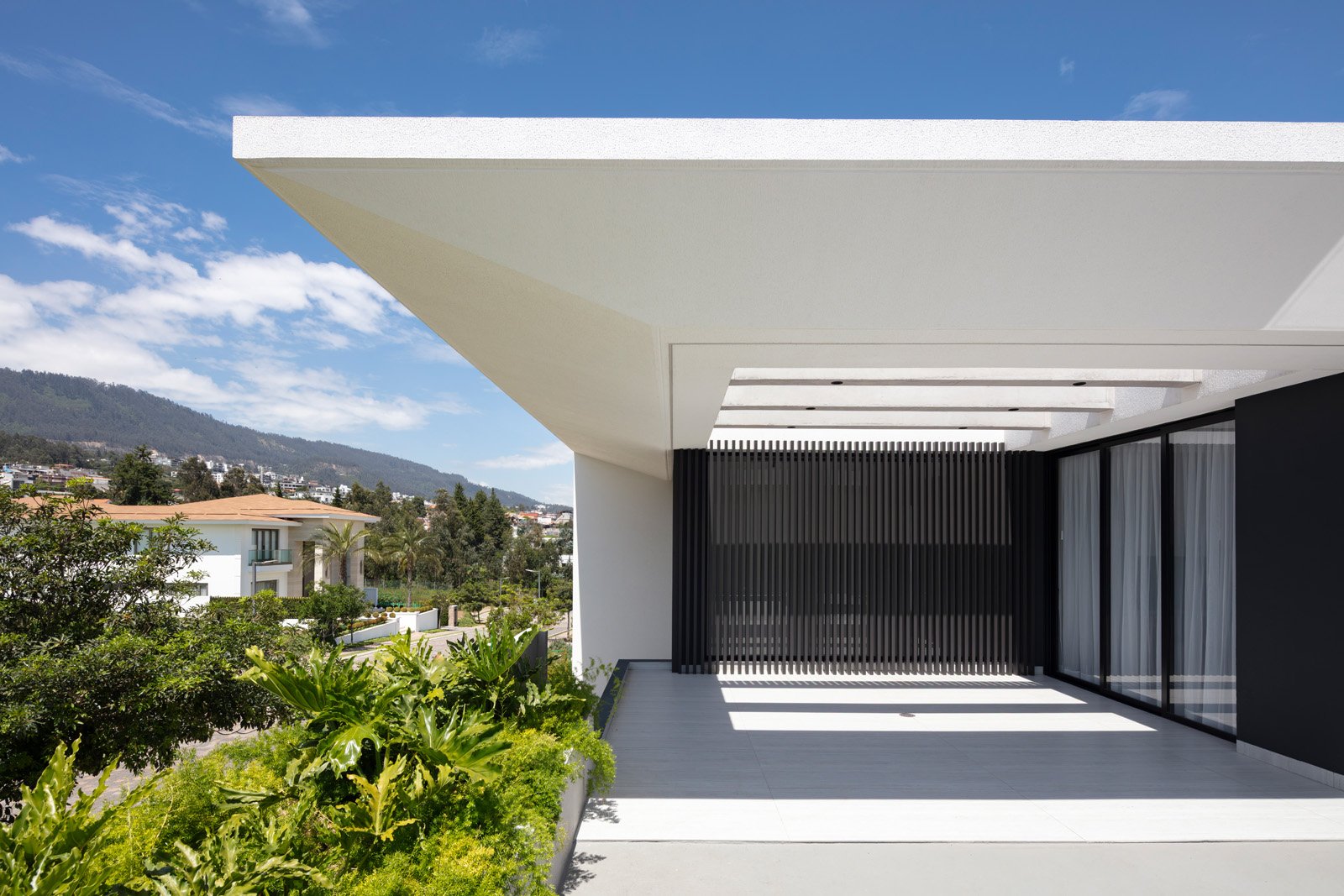 Vista de una terraza moderna con paredes blancas y negras, con ventanas grandes y cortinas transparentes, rodeada de vegetación, con una colina y casas en el fondo bajo un cielo azul con nubes.