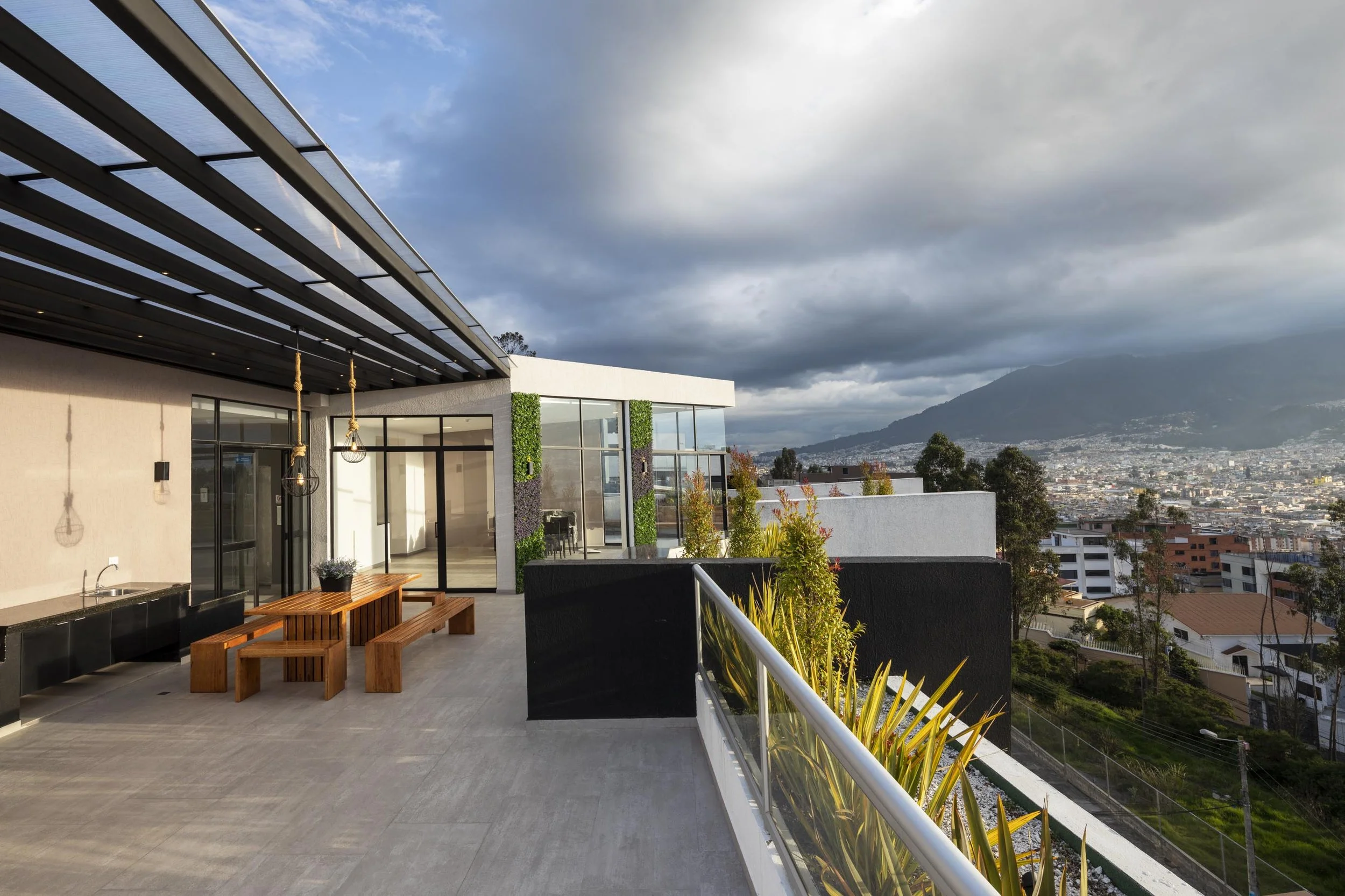 Amplia terraza de un edificio moderno con muebles de madera, jardineras y vista de la ciudad y montañas al fondo bajo un cielo nublado.