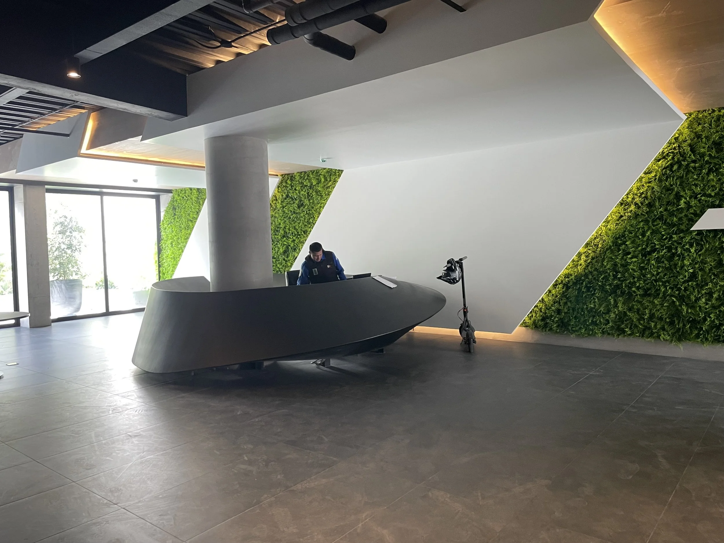 Recepción moderna con mostrador negro, un empleado en el fondo y paredes verdes decorativas. Ventanas grandes permiten entrada de luz natural y una bicicleta eléctrica está apoyada en la pared.