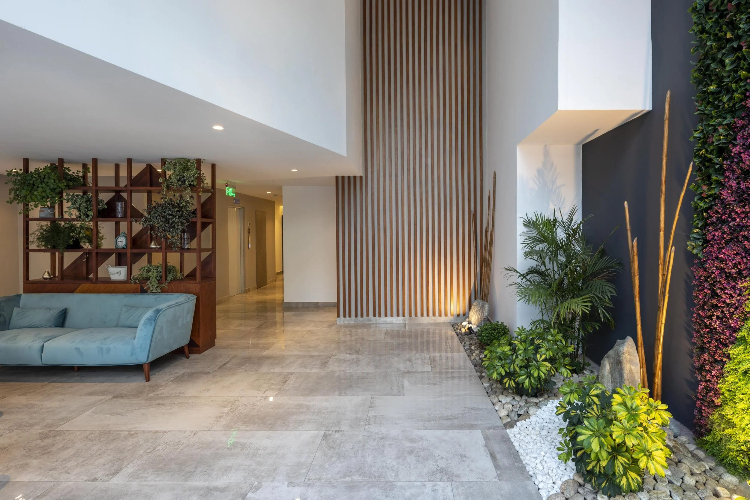 Vestíbulo decorativo con plantas y mueble de madera, paredes blancas y negras, suelo de baldosas y zona de descanso con sillón azul.